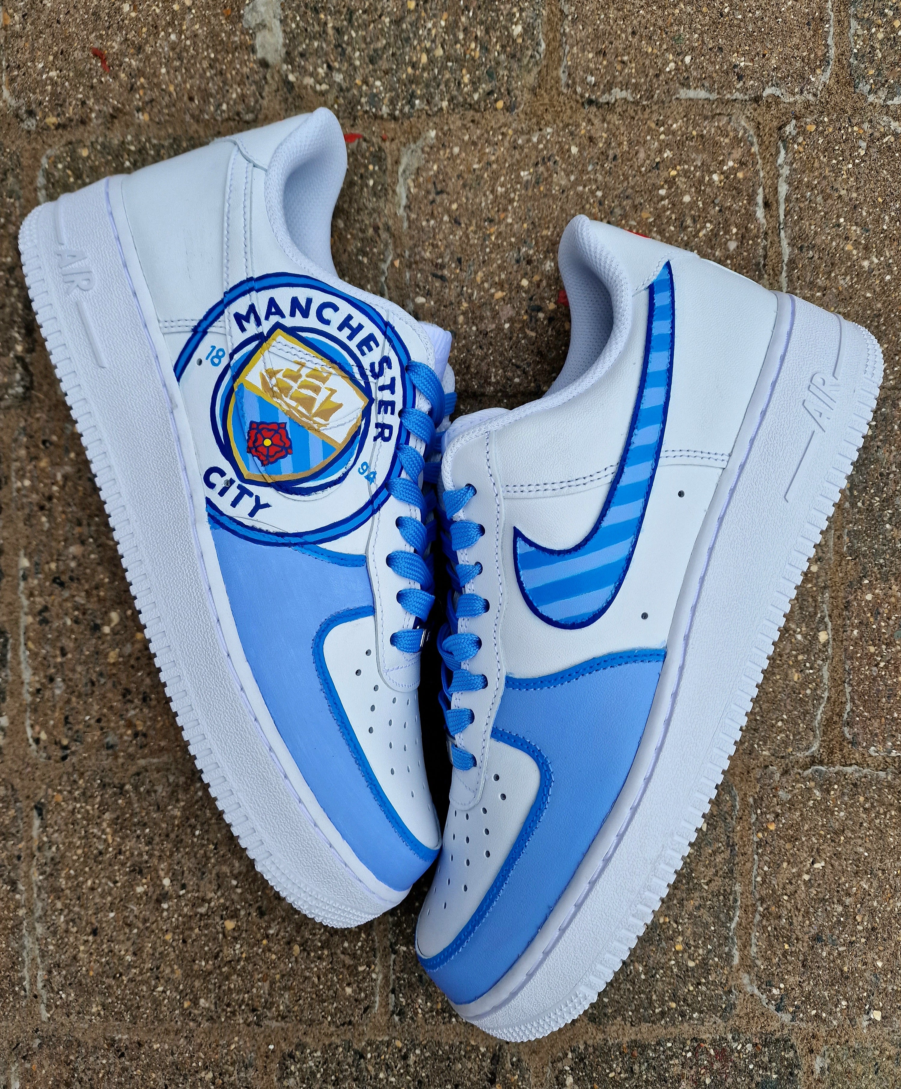 A pair of custom hand-painted Nike Air Force 1 sneakers featuring detailed artwork of the Manchester City Football Club crest, signature blue hues, striped swoosh, and personalized finishing, crafted by Gitartsy for football fans and sneaker collectors.

