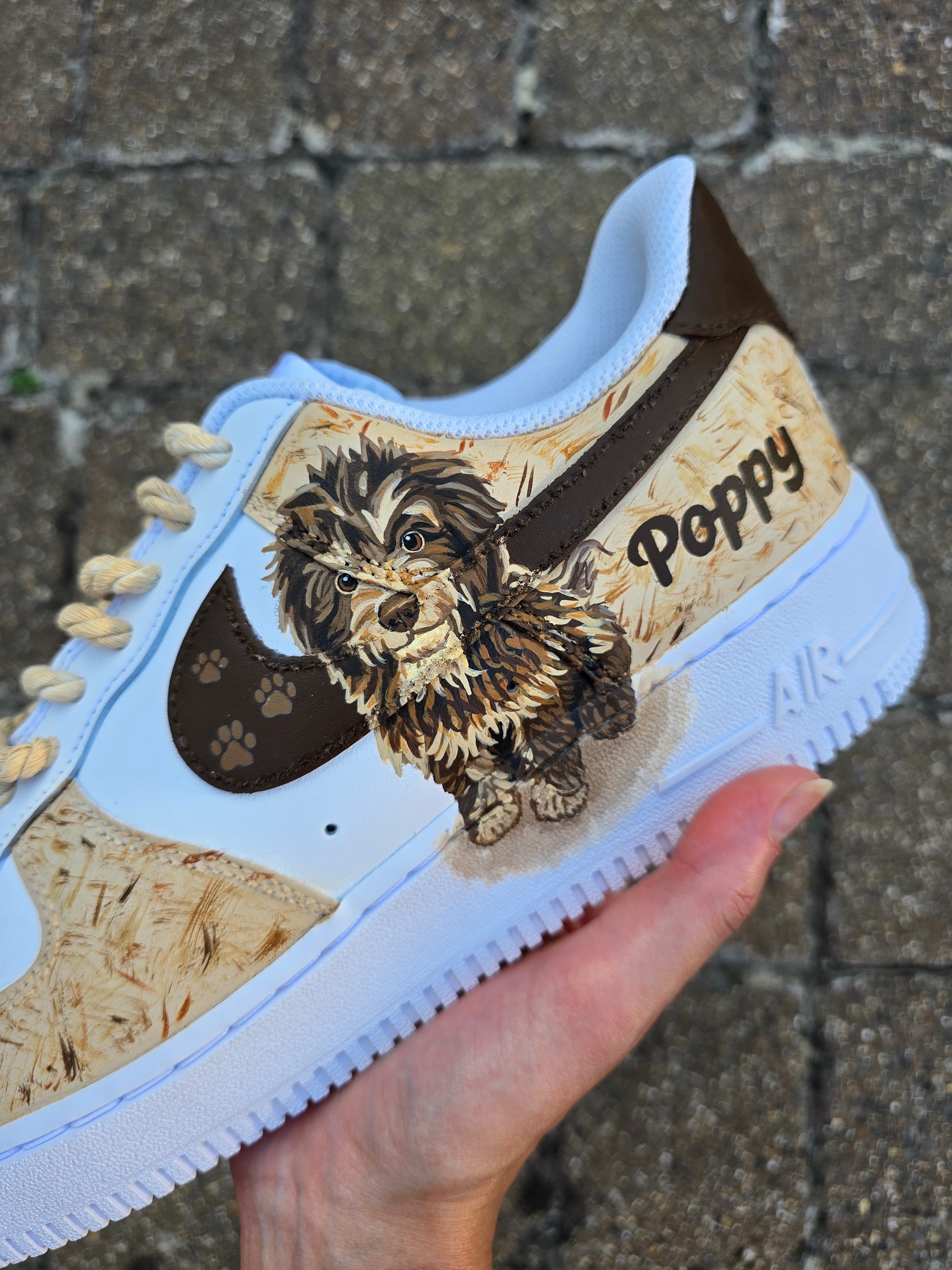 Custom hand-painted Nike Air Force 1 sneakers featuring a detailed portrait of a fluffy brown dog named Poppy 🐶. The shoes have a white leather base with light brown textured paint accents and a rich chocolate-brown swoosh adorned with paw prints 🐾. The pet’s name is written in a playful font, and tan rope laces add a rustic touch. Perfect for showcasing love for your pet in a stylish and artistic way.

