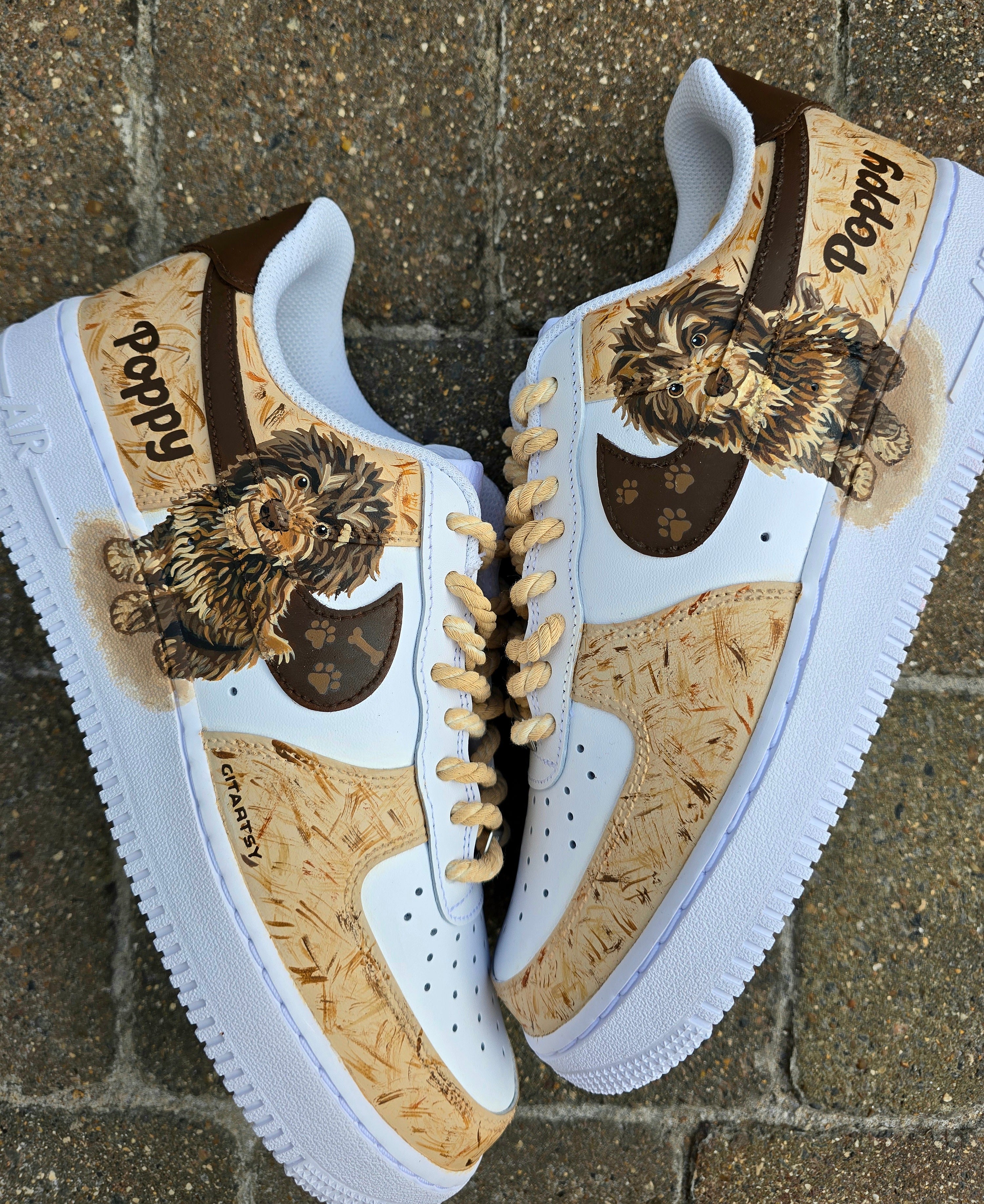 Custom hand-painted Nike Air Force 1 sneakers featuring a detailed portrait of a fluffy brown dog named Poppy 🐶. The shoes have a white leather base with light brown textured paint accents and a rich chocolate-brown swoosh adorned with paw prints 🐾. The pet’s name is written in a playful font, and tan rope laces add a rustic touch. Perfect for showcasing love for your pet in a stylish and artistic way.

