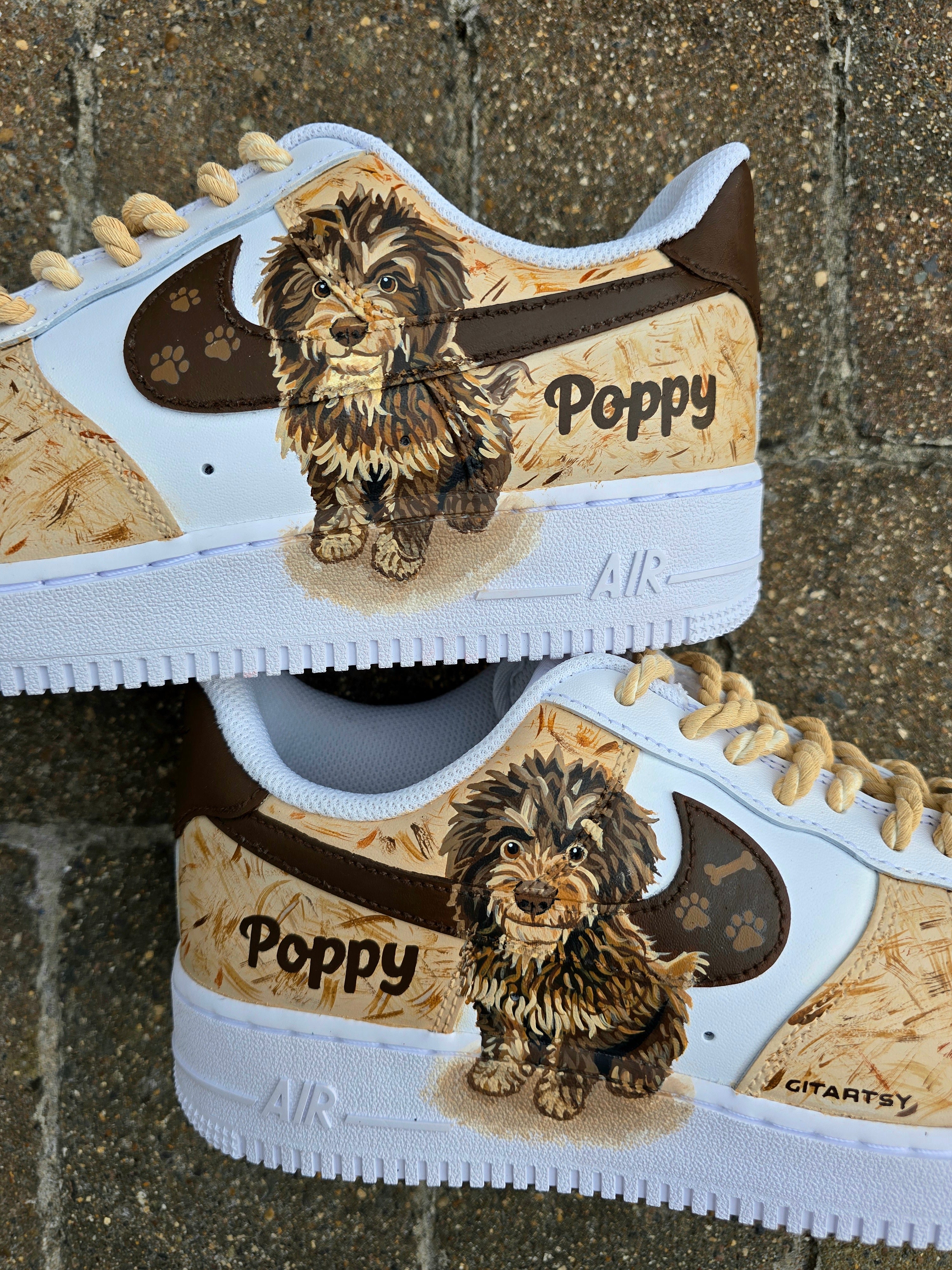 Custom hand-painted Nike Air Force 1 sneakers featuring a detailed portrait of a fluffy brown dog named Poppy 🐶. The shoes have a white leather base with light brown textured paint accents and a rich chocolate-brown swoosh adorned with paw prints 🐾. The pet’s name is written in a playful font, and tan rope laces add a rustic touch. Perfect for showcasing love for your pet in a stylish and artistic way.

