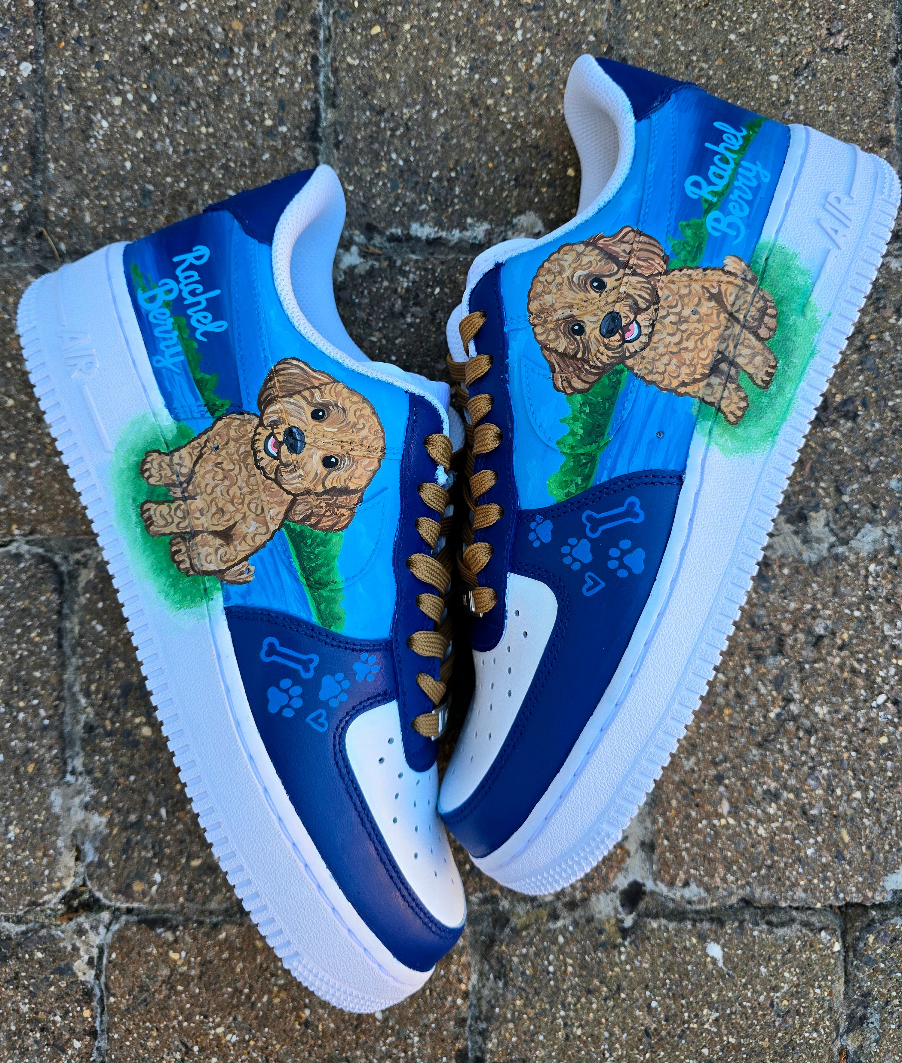 Custom Nike Air Force 1 sneakers 🐾 hand-painted with a detailed toy poodle pet portrait, featuring a caramel-colored curly-haired dog sitting in a grassy field with a blue sky and lake background. Includes the pet’s name in stylish script, paw prints, bones, and heart icons, all painted on a blue and navy color-block base with golden-brown laces. Each pair is personalized to match your pet’s likeness and crafted using durable, waterproof leather paints, making them wearable art for dog lovers.

