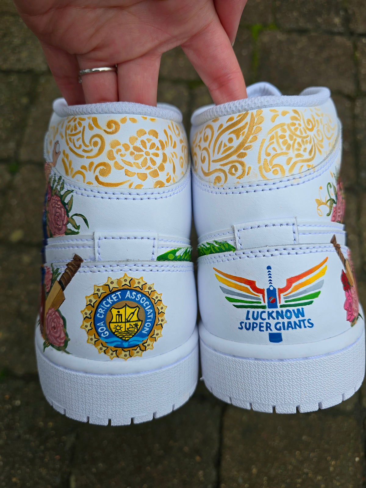 A pair of nike jordan 1 white high-top sneakers with custom illustrations of a couple Arjun Tendulkar and Saaniya Chandhok with gold paisley patterns henna, pets, florals with maroon and blue colour scheme displayed against a dark red background with a GITARTSY logo in the corner. Cricket team logos logos on the backs