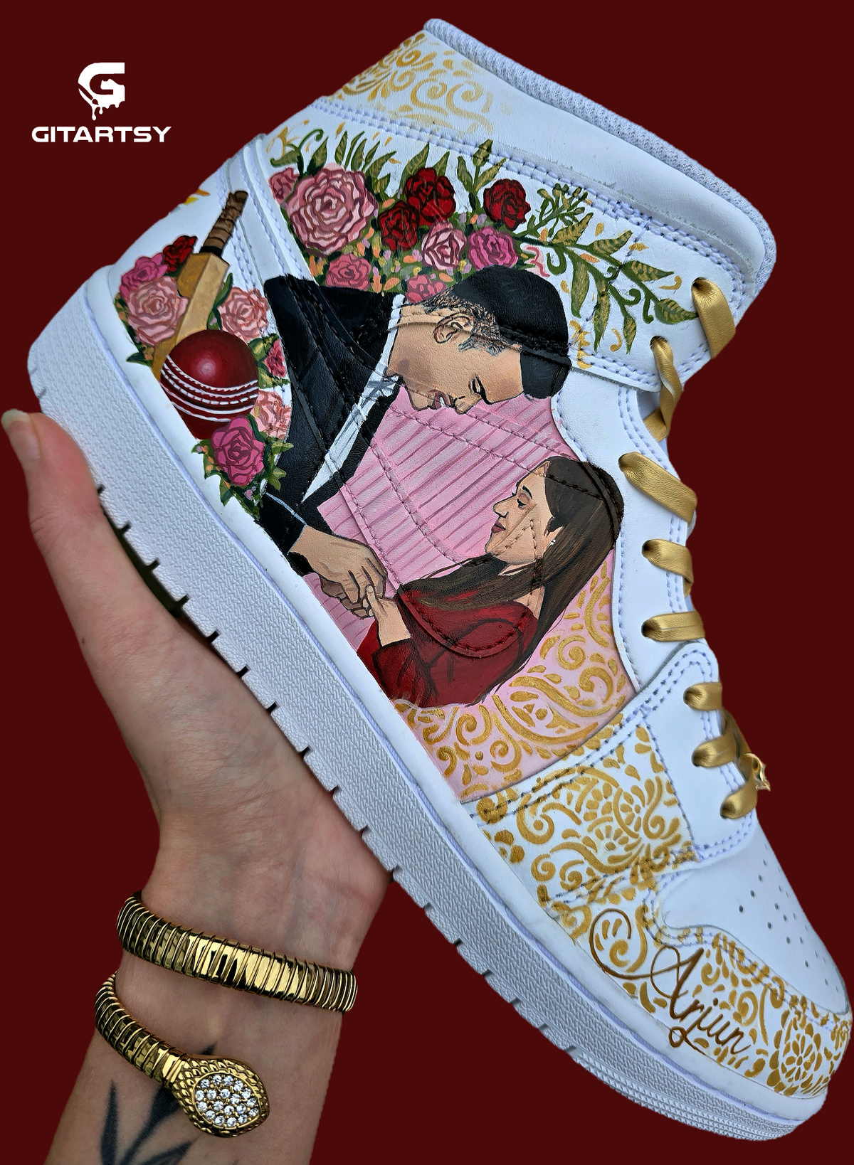 A pair of nike jordan 1 white high-top sneakers with custom illustrations of a couple Arjun Tendulkar and Saaniya Chandhok with gold paisley patterns henna, pets, florals with maroon and blue colour scheme displayed against a dark red background with a GITARTSY logo in the corner.