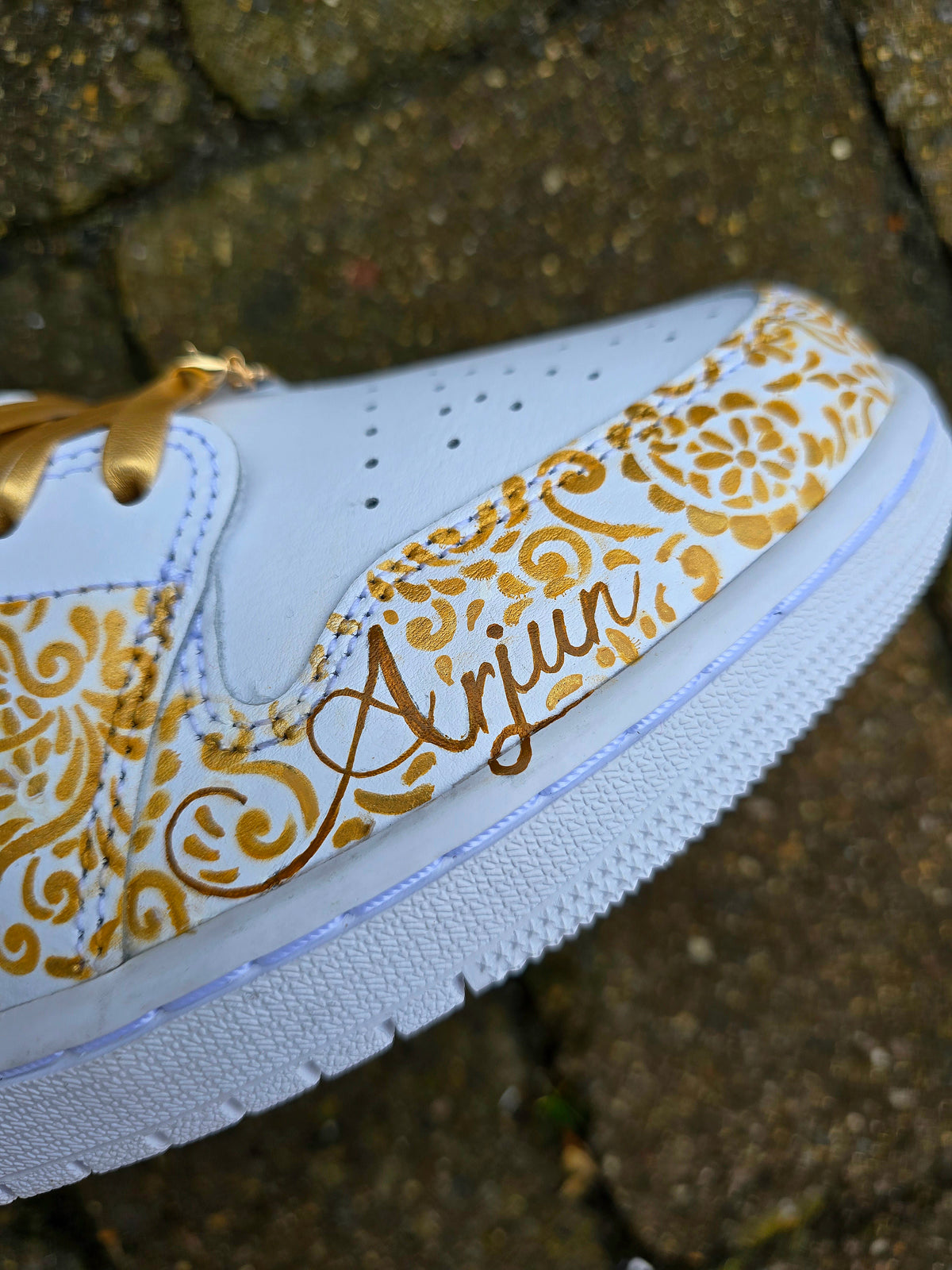 A close-up of a white sneaker with gold floral scroll patterns and the name 'Arjun' written in gold script on the side, photographed outdoors against a stone surface.