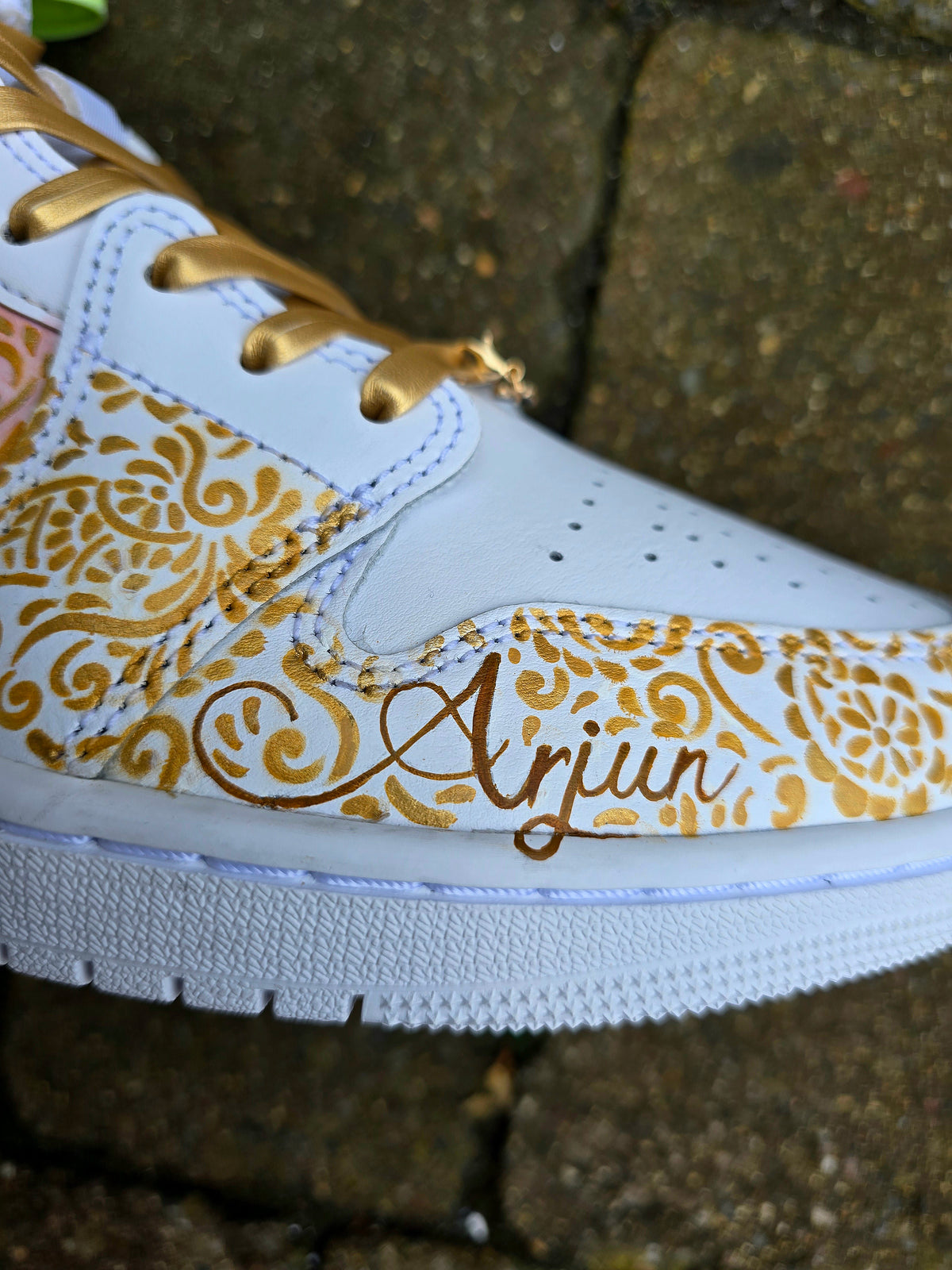 A close-up of a white sneaker with gold floral patterns and the name 'Anjun' written in gold script on the side, photographed outdoors against a stone background.