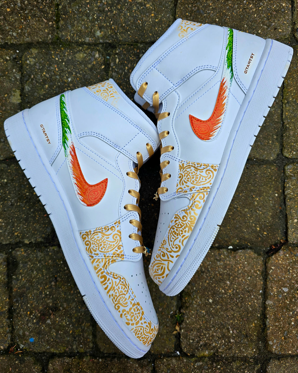 A pair of nike jordan 1 white high-top sneakers with custom illustrations of a couple Arjun Tendulkar and Saaniya Chandhok with gold paisley patterns henna, pets, florals with maroon and blue colour scheme displayed against a dark red background with a GITARTSY logo in the corner.