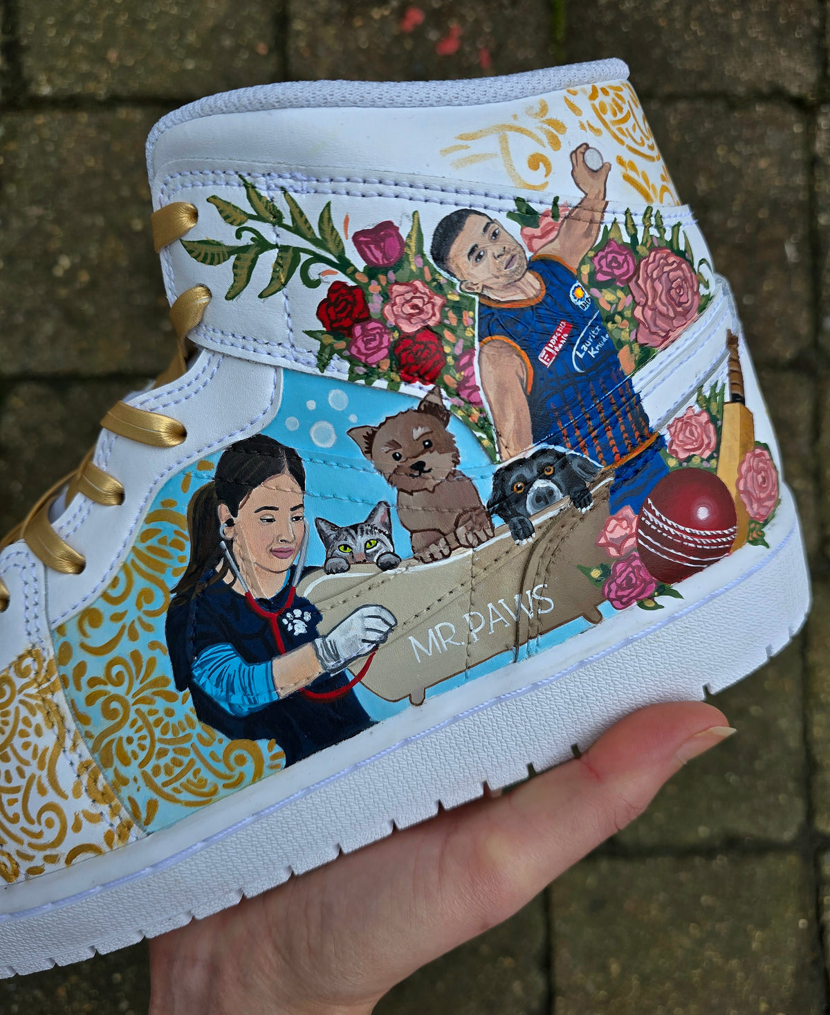 A pair of nike jordan 1 white high-top sneakers with custom illustrations of a couple Arjun Tendulkar and Saaniya Chandhok with gold paisley patterns henna, pets, florals with maroon and blue colour scheme displayed against a dark red background with a GITARTSY logo in the corner.