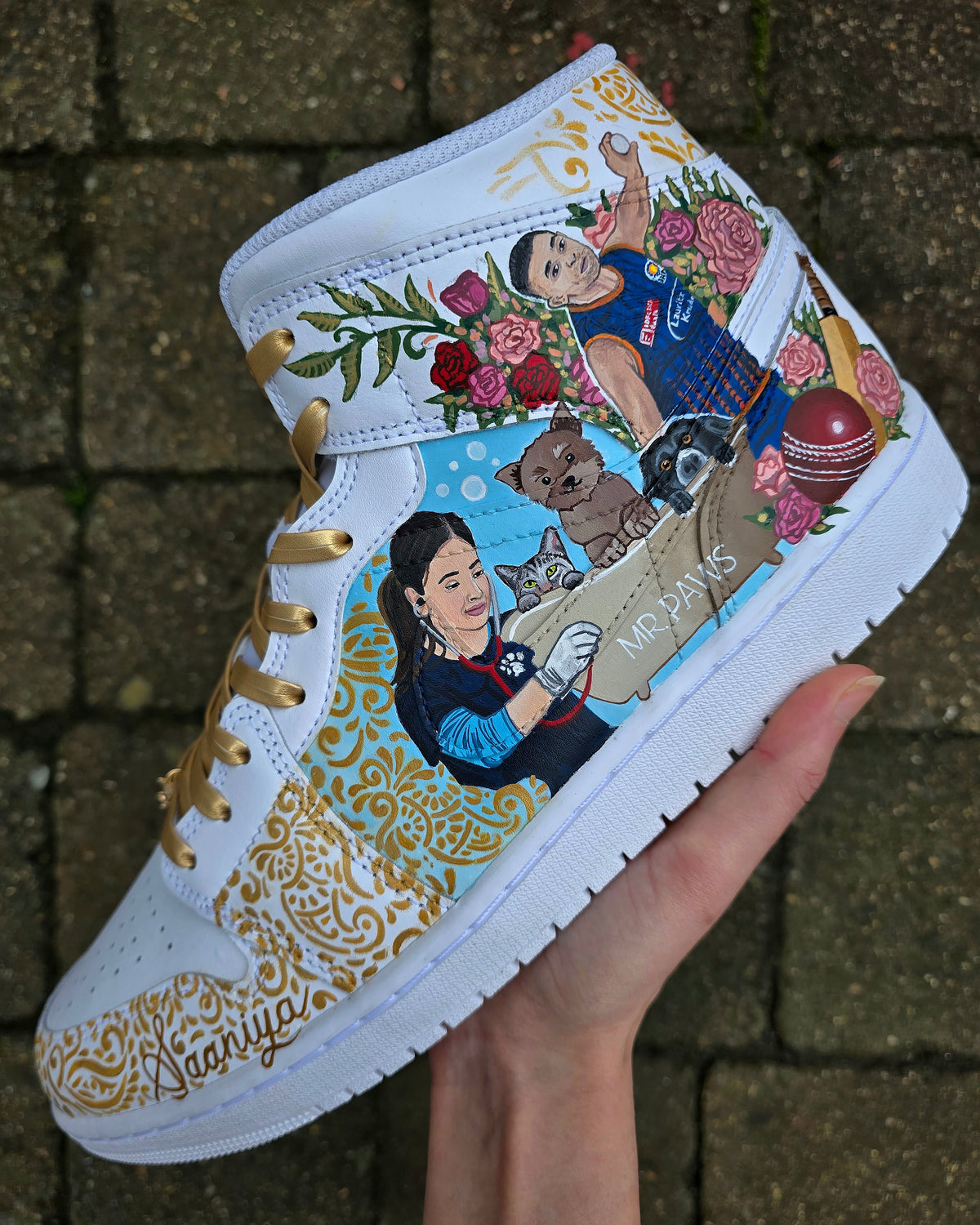 A pair of nike jordan 1 white high-top sneakers with custom illustrations of a couple Arjun Tendulkar and Saaniya Chandhok with gold paisley patterns henna, pets, florals with maroon and blue colour scheme displayed against a dark red background with a GITARTSY logo in the corner.