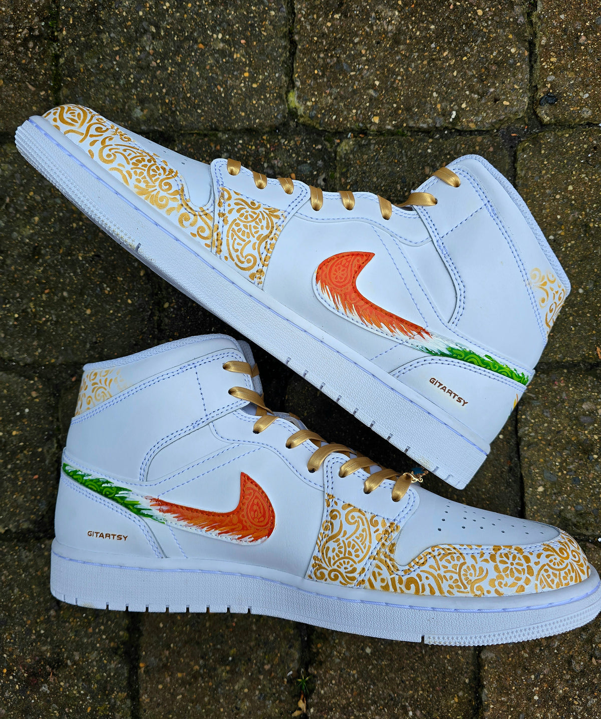 A pair of nike jordan 1 white high-top sneakers with custom illustrations of a couple Arjun Tendulkar and Saaniya Chandhok with gold paisley patterns henna, pets, florals with maroon and blue colour scheme displayed against a dark red background with a GITARTSY logo in the corner.