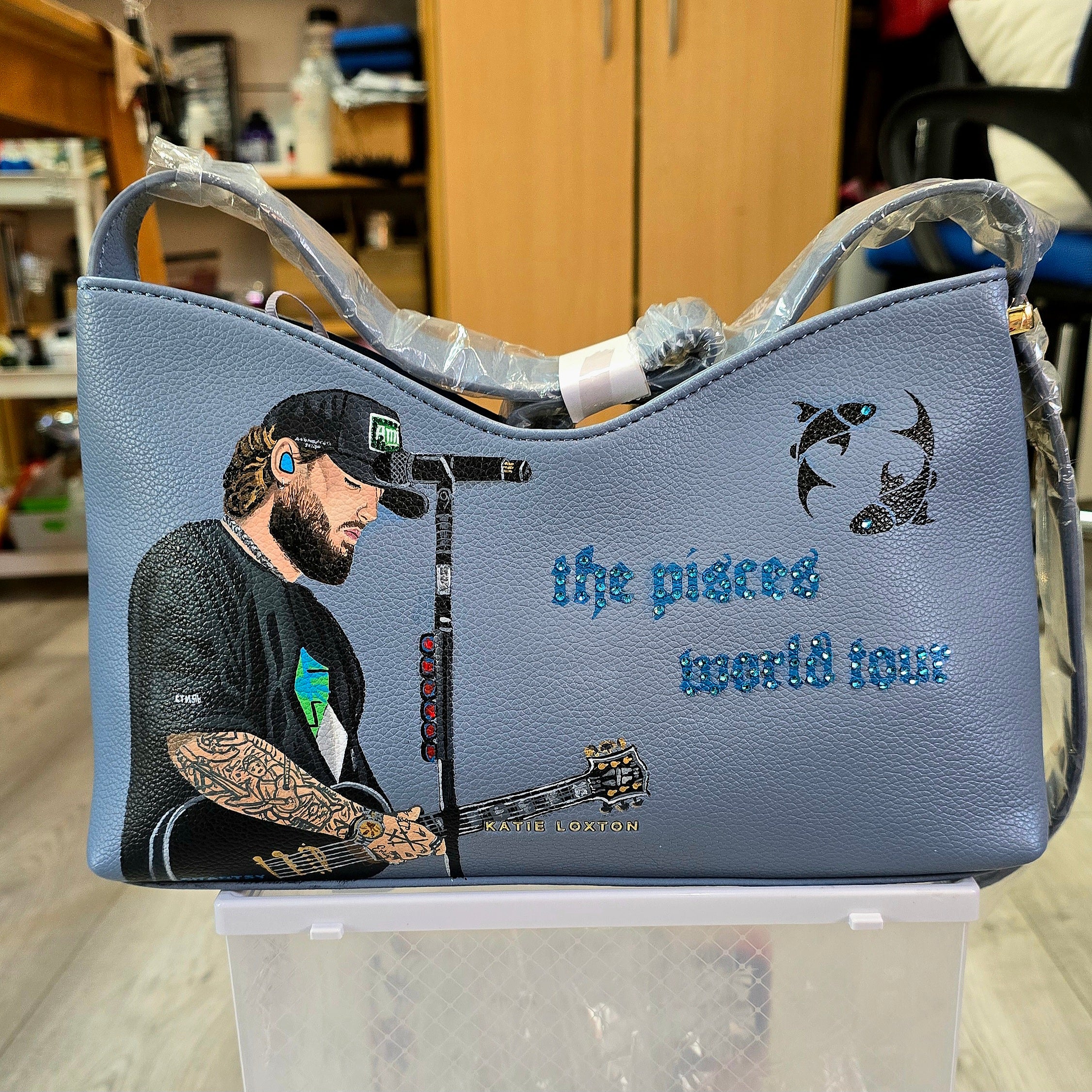 Hand-painted vegan leather handbag featuring highly detailed artwork of a musical artist performing, enhanced with rhinestone embellishments, microphone details, tattoos, and vibrant accessories, all painted on a soft leather-textured blue surface. The bag is styled with a structured handle, protective plastic wrap, and is ideal for fans and fashion-forward individuals who appreciate handcrafted accessories.

