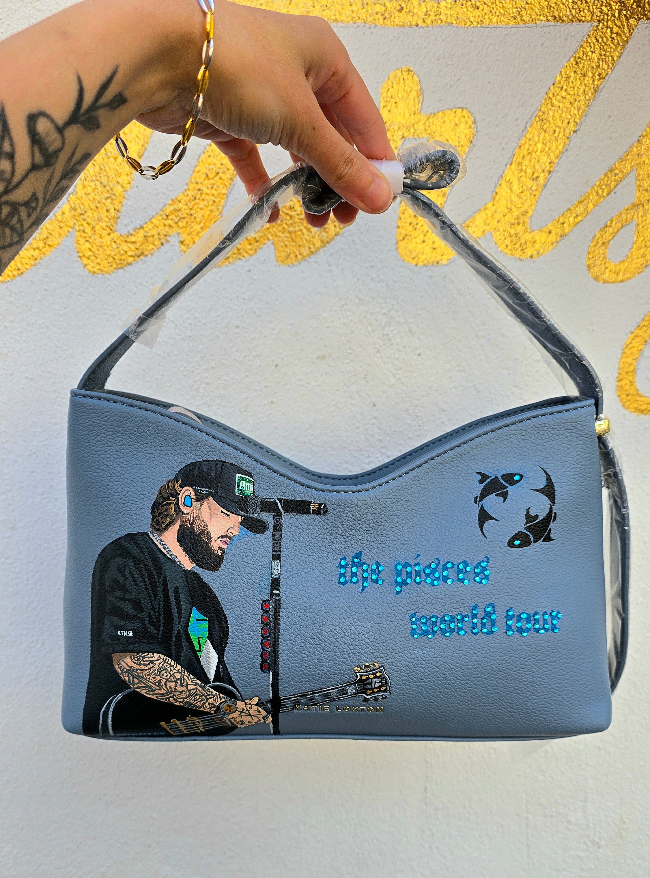 Hand-painted vegan leather handbag featuring highly detailed artwork of a musical artist performing, enhanced with rhinestone embellishments, microphone details, tattoos, and vibrant accessories, all painted on a soft leather-textured blue surface. The bag is styled with a structured handle, protective plastic wrap, and is ideal for fans and fashion-forward individuals who appreciate handcrafted accessories.

