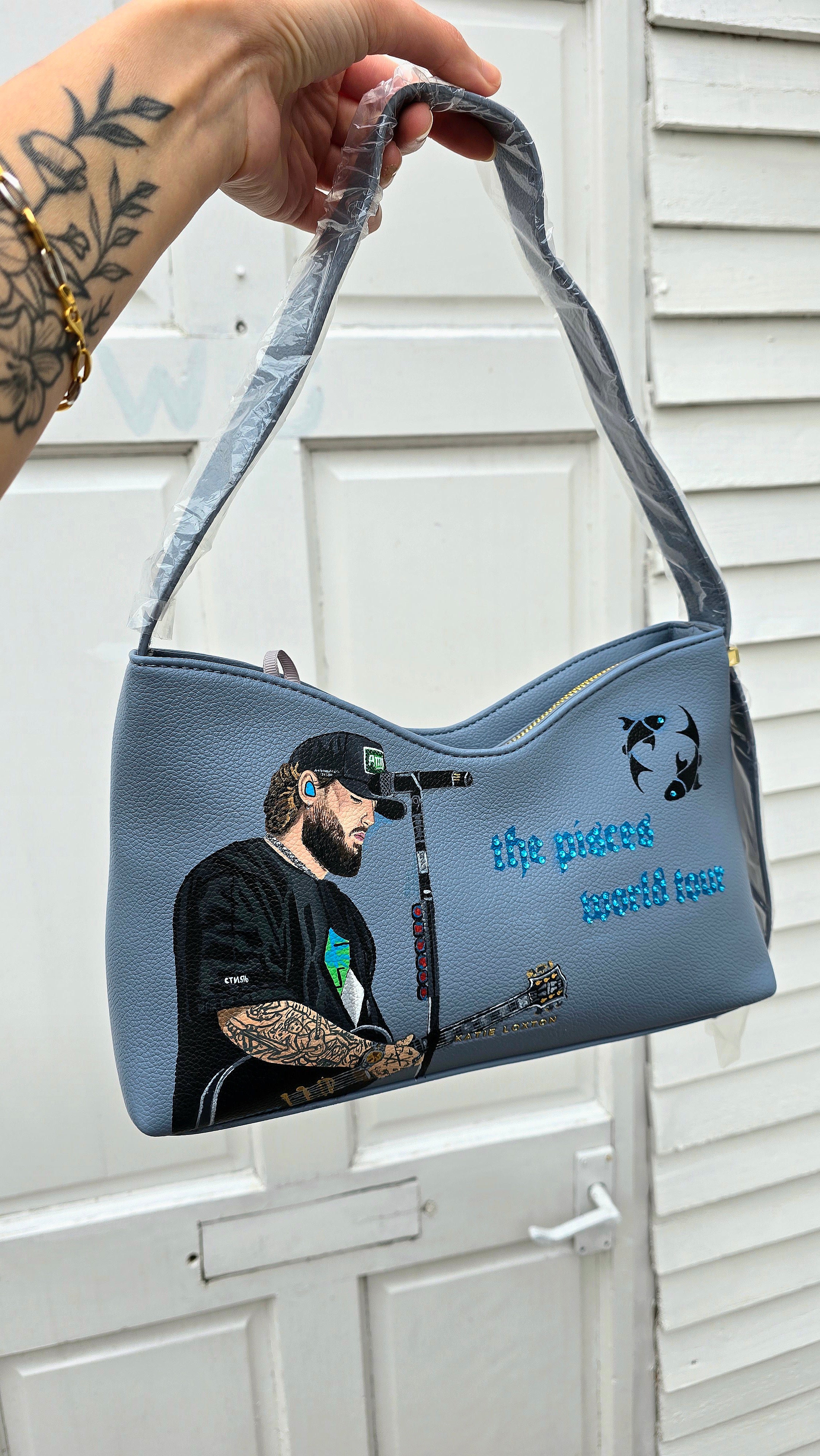 Hand-painted vegan leather handbag featuring highly detailed artwork of a musical artist performing, enhanced with rhinestone embellishments, microphone details, tattoos, and vibrant accessories, all painted on a soft leather-textured blue surface. The bag is styled with a structured handle, protective plastic wrap, and is ideal for fans and fashion-forward individuals who appreciate handcrafted accessories.

