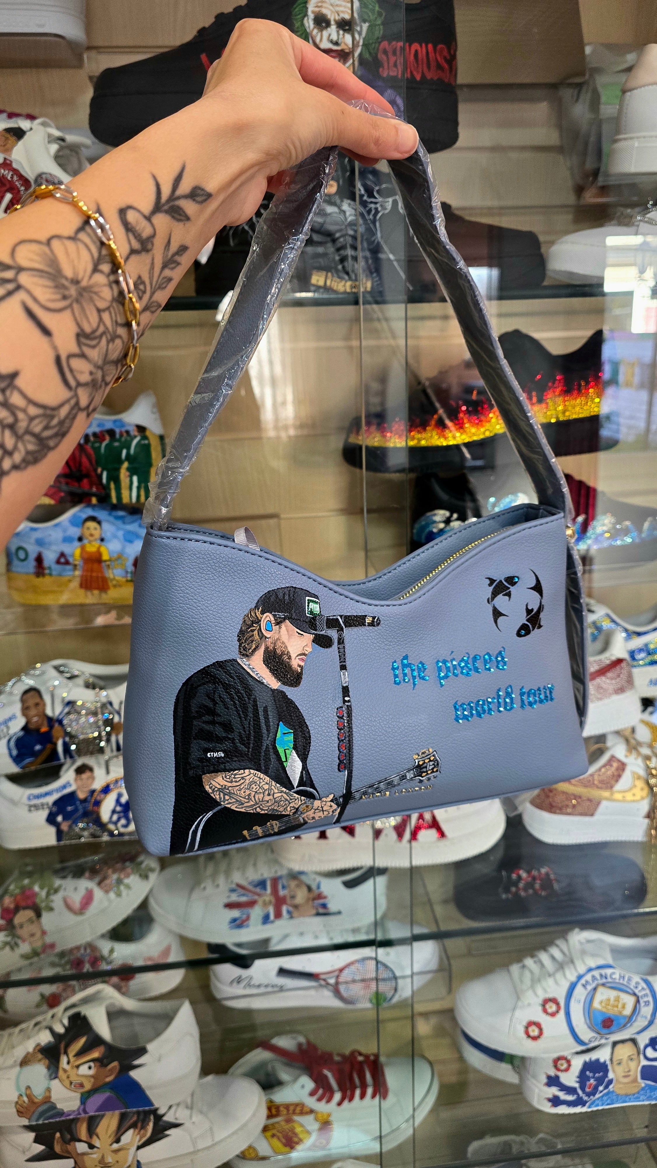 Hand-painted vegan leather handbag featuring highly detailed artwork of a musical artist performing, enhanced with rhinestone embellishments, microphone details, tattoos, and vibrant accessories, all painted on a soft leather-textured blue surface. The bag is styled with a structured handle, protective plastic wrap, and is ideal for fans and fashion-forward individuals who appreciate handcrafted accessories.

