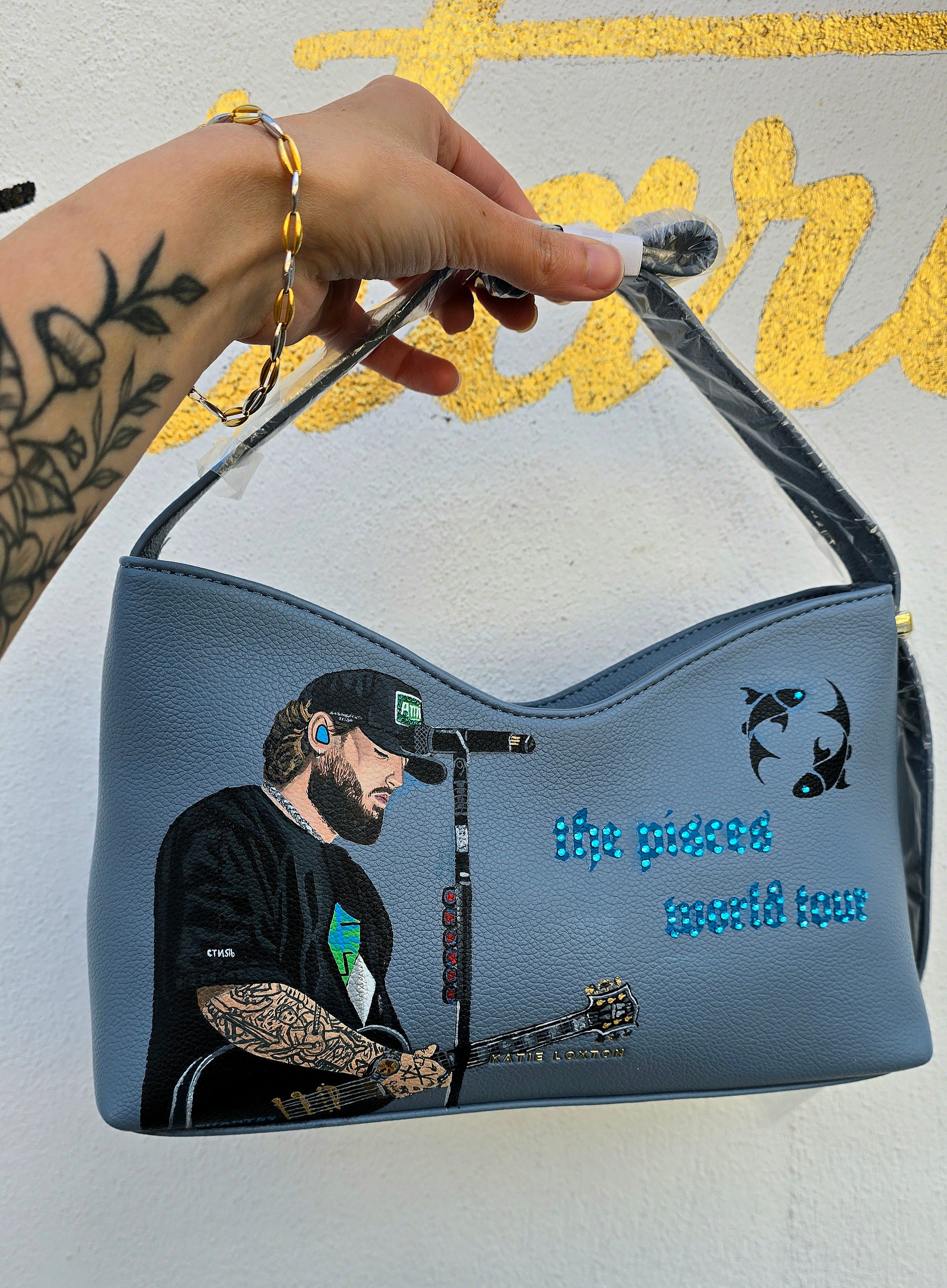 Hand-painted vegan leather handbag featuring highly detailed artwork of a musical artist performing, enhanced with rhinestone embellishments, microphone details, tattoos, and vibrant accessories, all painted on a soft leather-textured blue surface. The bag is styled with a structured handle, protective plastic wrap, and is ideal for fans and fashion-forward individuals who appreciate handcrafted accessories.

