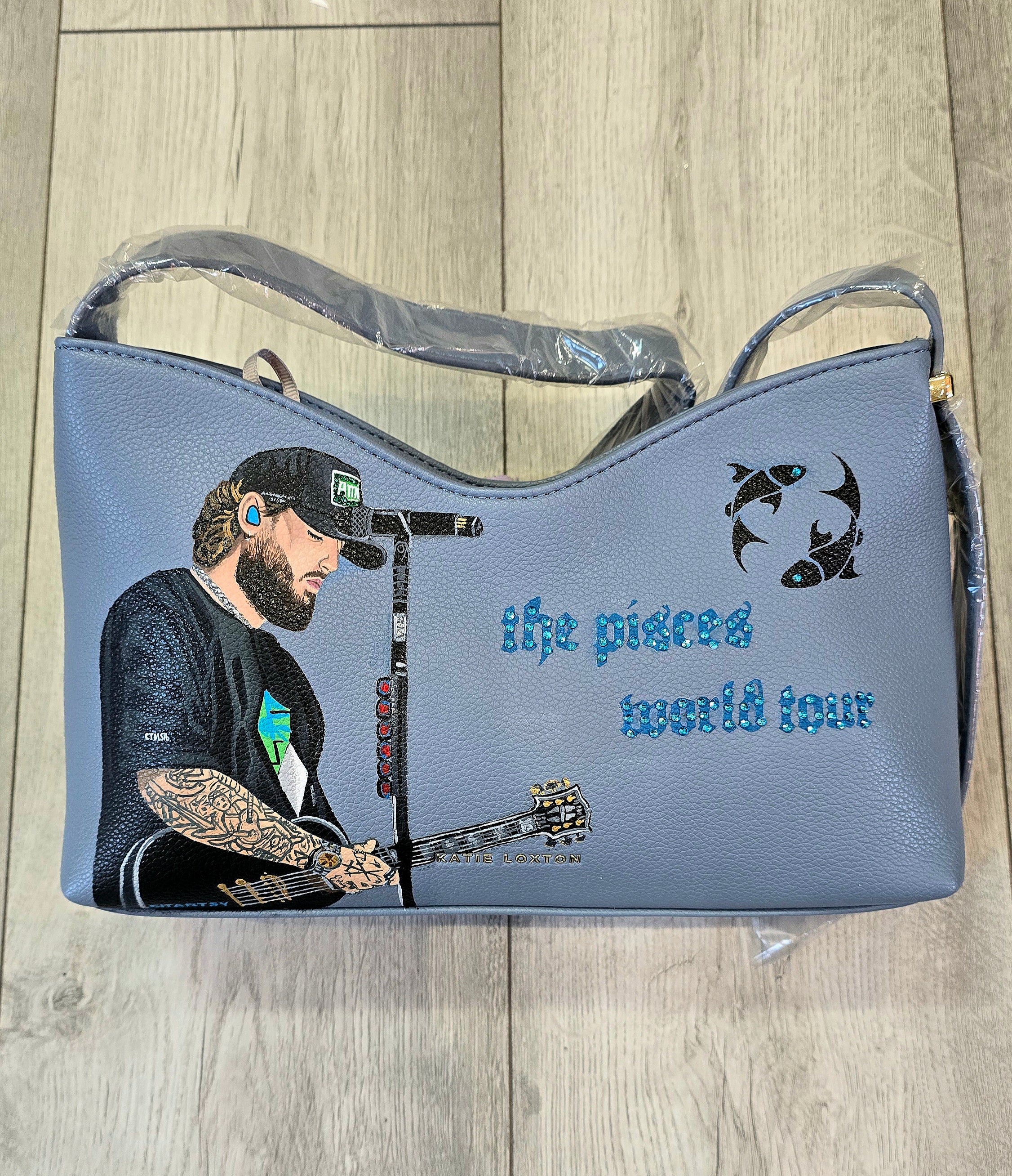 Hand-painted vegan leather handbag featuring highly detailed artwork of a musical artist performing, enhanced with rhinestone embellishments, microphone details, tattoos, and vibrant accessories, all painted on a soft leather-textured blue surface. The bag is styled with a structured handle, protective plastic wrap, and is ideal for fans and fashion-forward individuals who appreciate handcrafted accessories.


