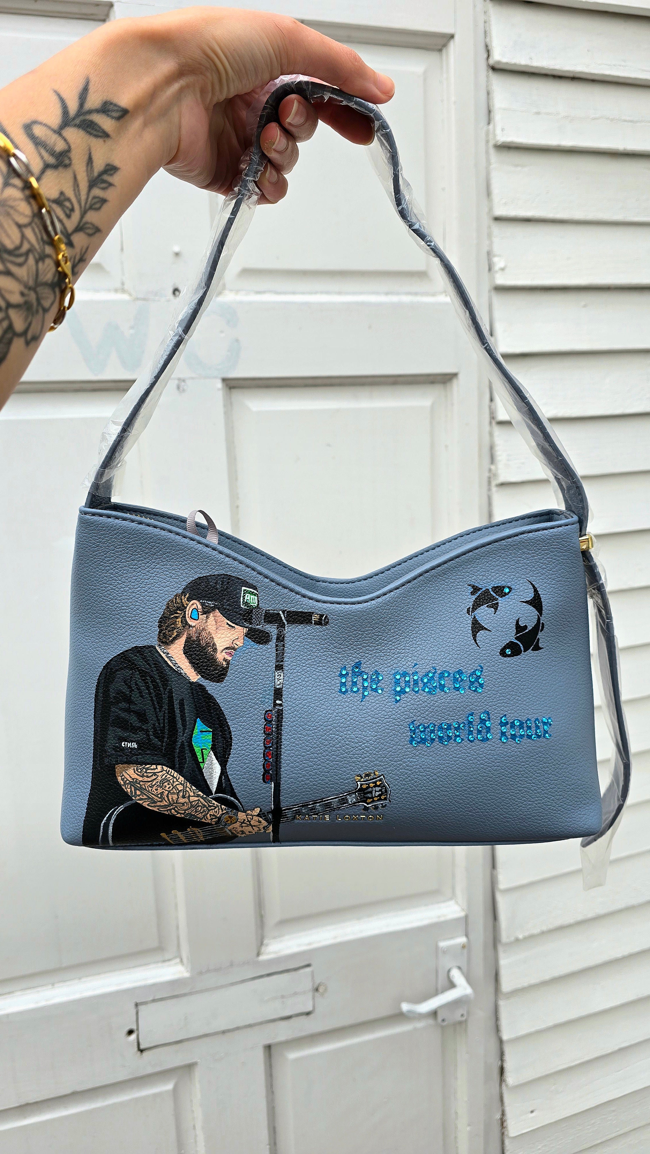 Hand-painted vegan leather handbag featuring highly detailed artwork of a musical artist performing, enhanced with rhinestone embellishments, microphone details, tattoos, and vibrant accessories, all painted on a soft leather-textured blue surface. The bag is styled with a structured handle, protective plastic wrap, and is ideal for fans and fashion-forward individuals who appreciate handcrafted accessories.

