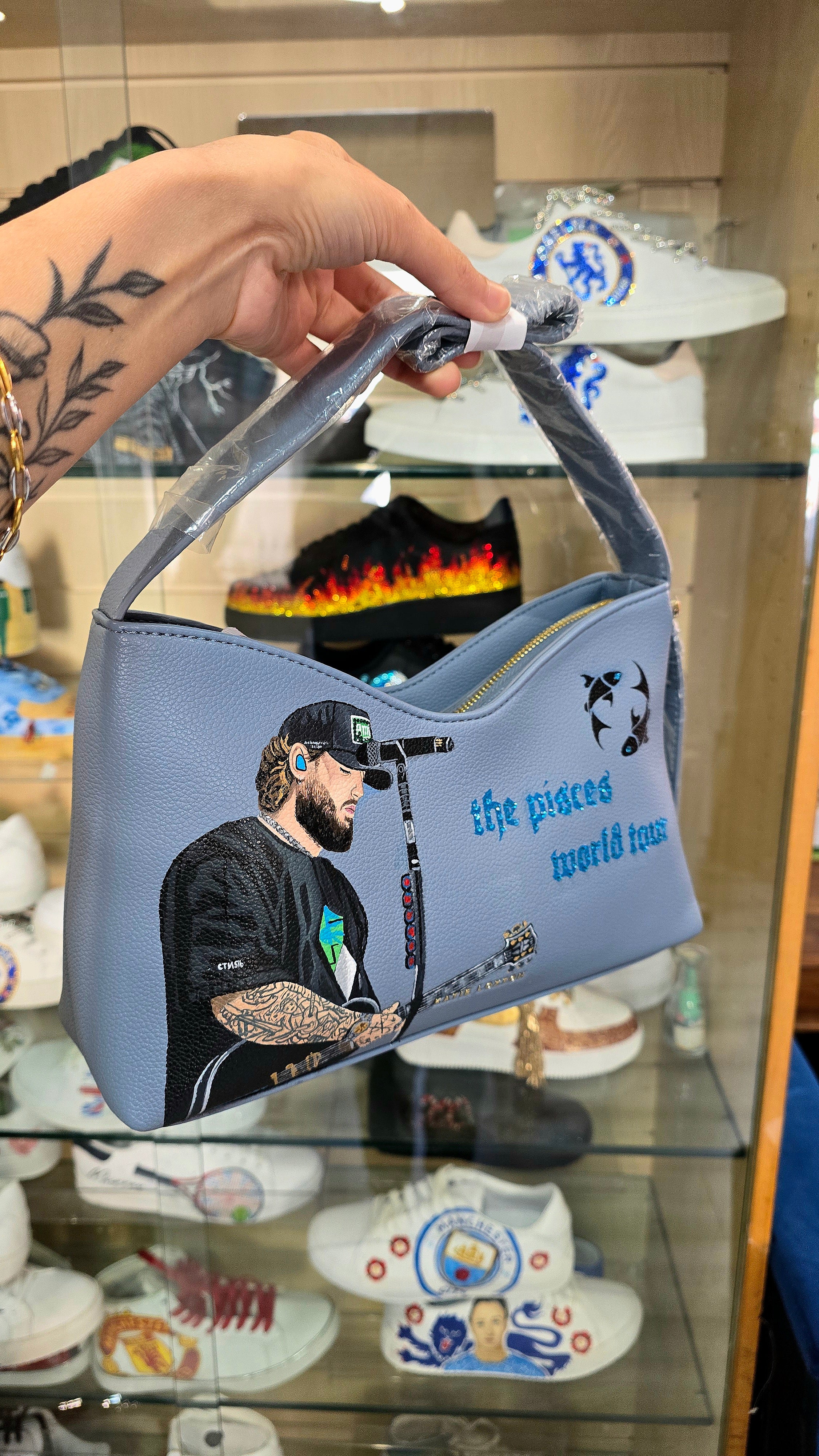 Hand-painted vegan leather handbag featuring highly detailed artwork of a musical artist performing, enhanced with rhinestone embellishments, microphone details, tattoos, and vibrant accessories, all painted on a soft leather-textured blue surface. The bag is styled with a structured handle, protective plastic wrap, and is ideal for fans and fashion-forward individuals who appreciate handcrafted accessories.

