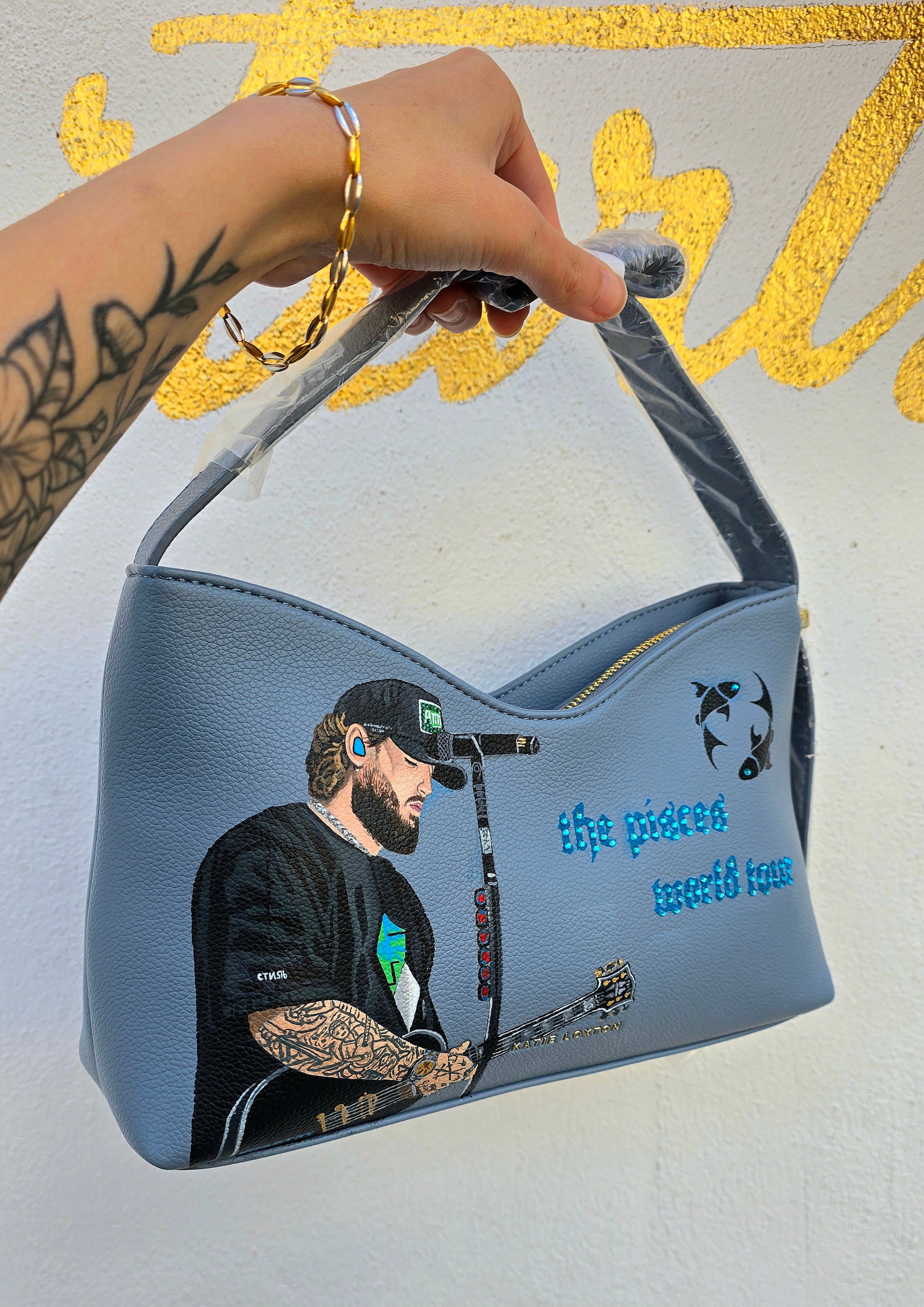 Hand-painted vegan leather handbag featuring highly detailed artwork of a musical artist performing, enhanced with rhinestone embellishments, microphone details, tattoos, and vibrant accessories, all painted on a soft leather-textured blue surface. The bag is styled with a structured handle, protective plastic wrap, and is ideal for fans and fashion-forward individuals who appreciate handcrafted accessories.


