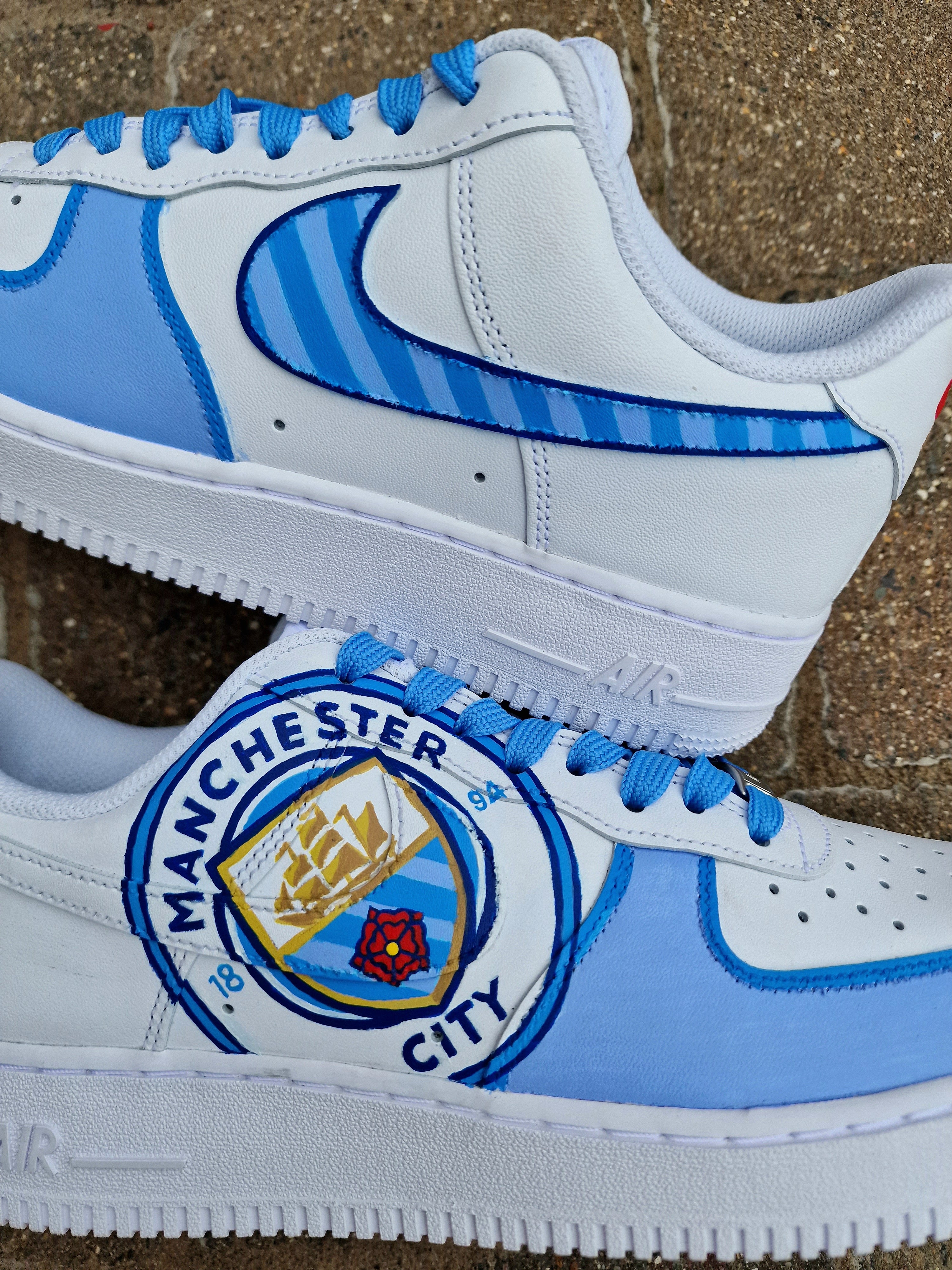 A pair of custom hand-painted Nike Air Force 1 sneakers featuring detailed artwork of the Manchester City Football Club crest, signature blue hues, striped swoosh, and personalized finishing, crafted by Gitartsy for football fans and sneaker collectors.

