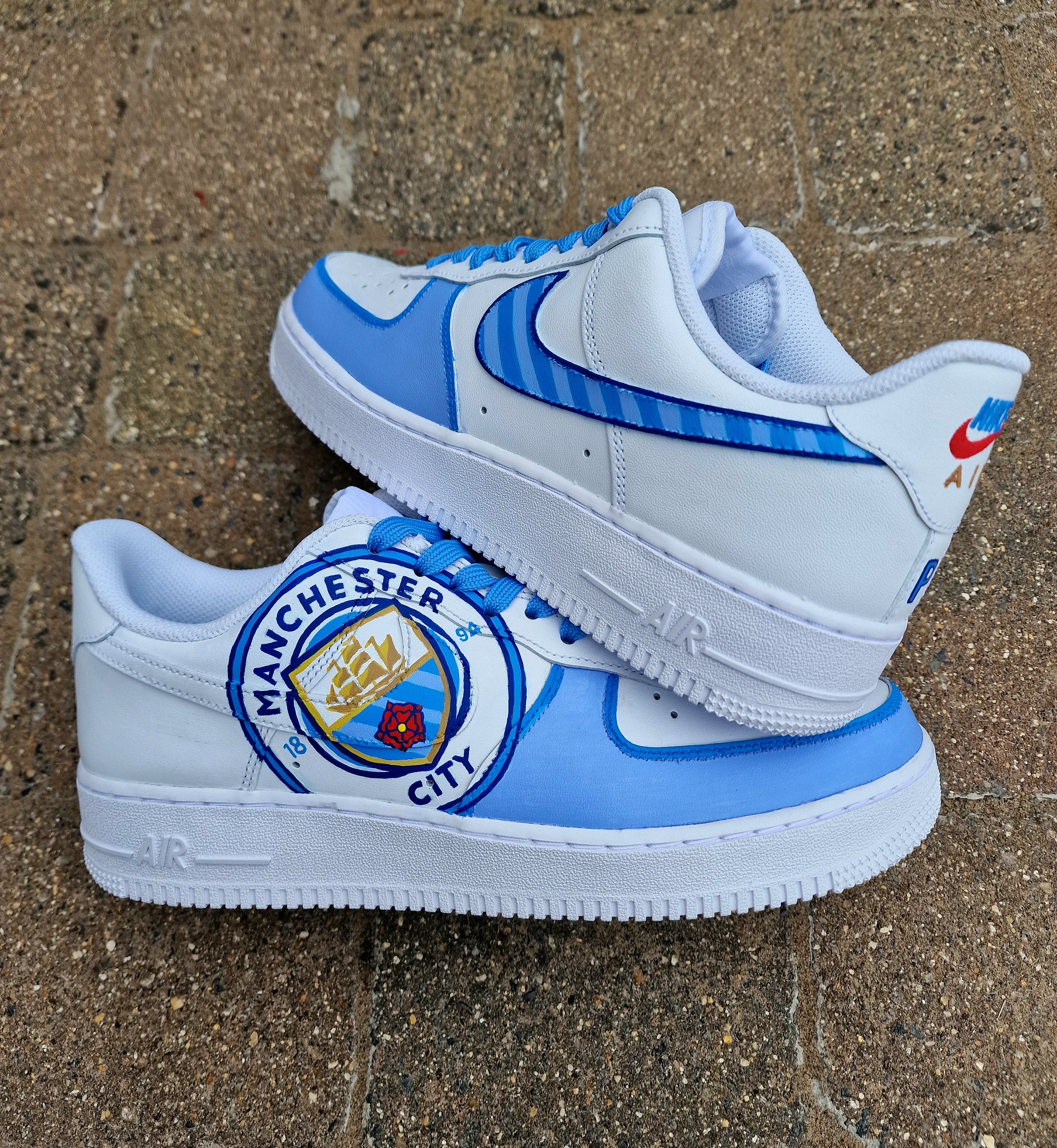 A pair of custom hand-painted Nike Air Force 1 sneakers featuring detailed artwork of the Manchester City Football Club crest, signature blue hues, striped swoosh, and personalized finishing, crafted by Gitartsy for football fans and sneaker collectors.

