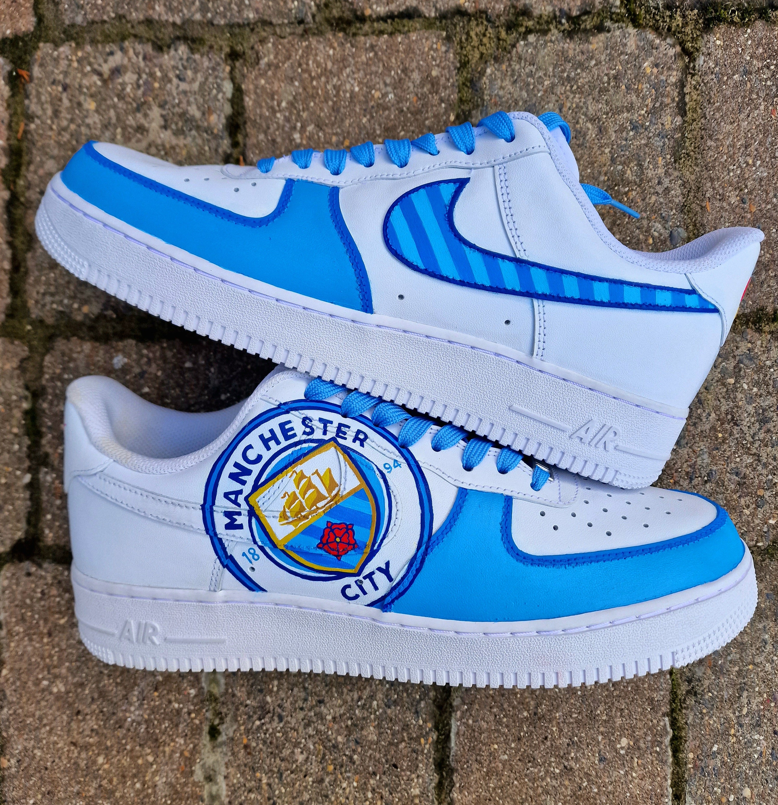 A pair of custom hand-painted Nike Air Force 1 sneakers featuring detailed artwork of the Manchester City Football Club crest, signature blue hues, striped swoosh, and personalized finishing, crafted by Gitartsy for football fans and sneaker collectors.

