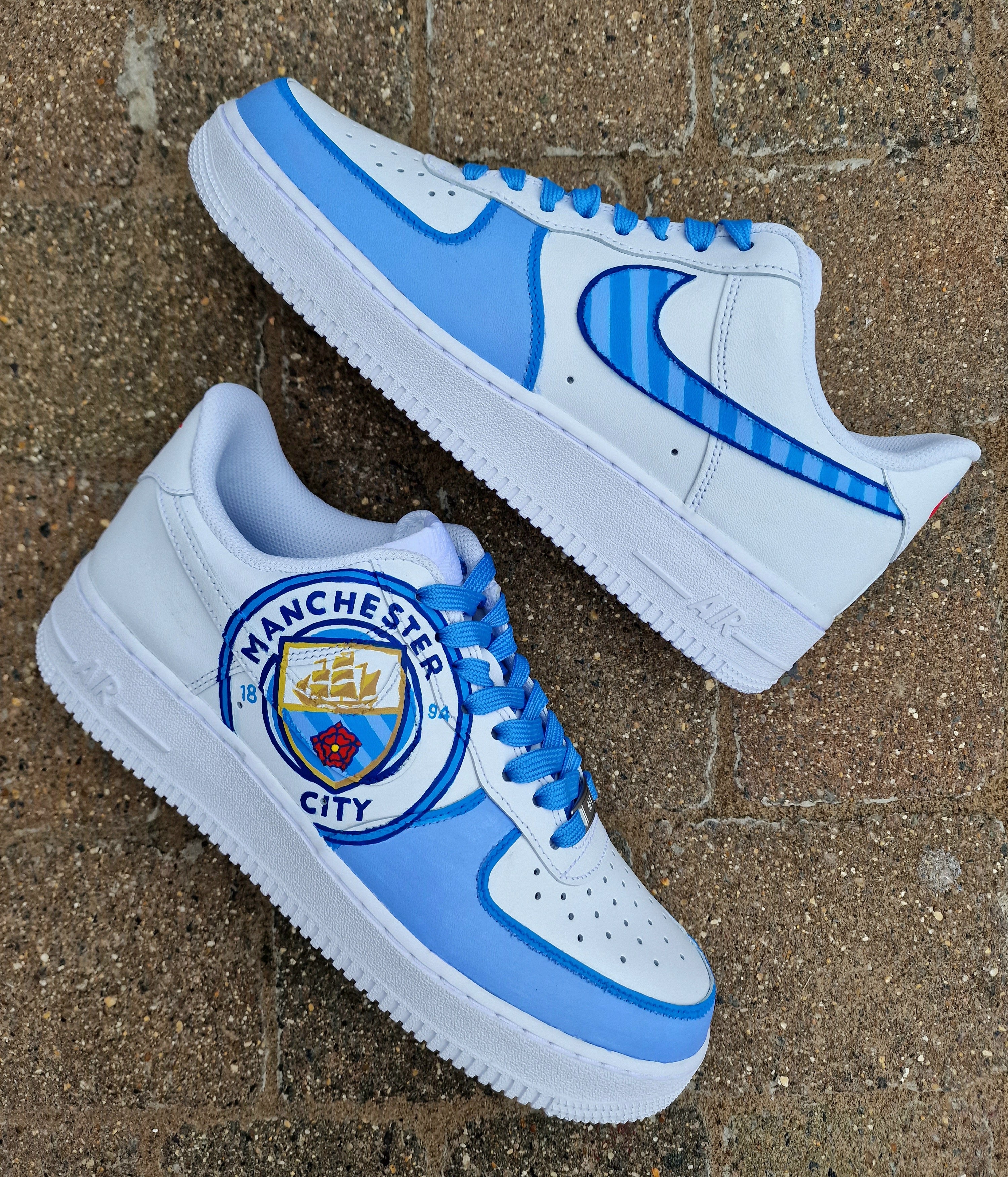 A pair of custom hand-painted Nike Air Force 1 sneakers featuring detailed artwork of the Manchester City Football Club crest, signature blue hues, striped swoosh, and personalized finishing, crafted by Gitartsy for football fans and sneaker collectors.

