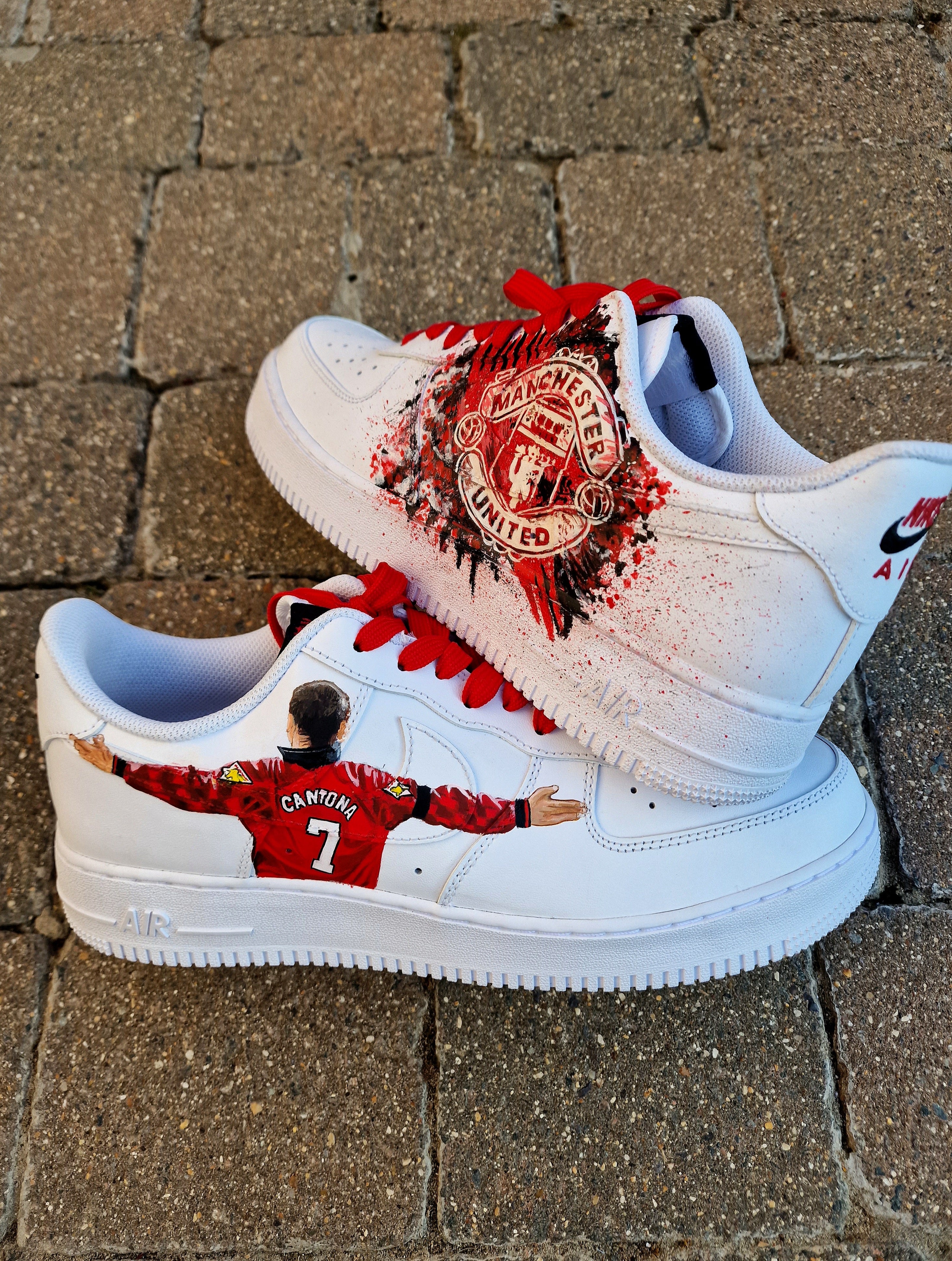 Custom Nike Air Force 1 sneakers hand-painted with Manchester United's club crest on one side in bold red-black splatter, and Eric Cantona's famous goal celebration pose with arms outstretched wearing the No. 7 jersey on the opposite side. Finished with red laces and premium leather base, these AF1s are waterproof-coated and crafted with attention to detail, offering a perfect gift for Manchester United fans.

