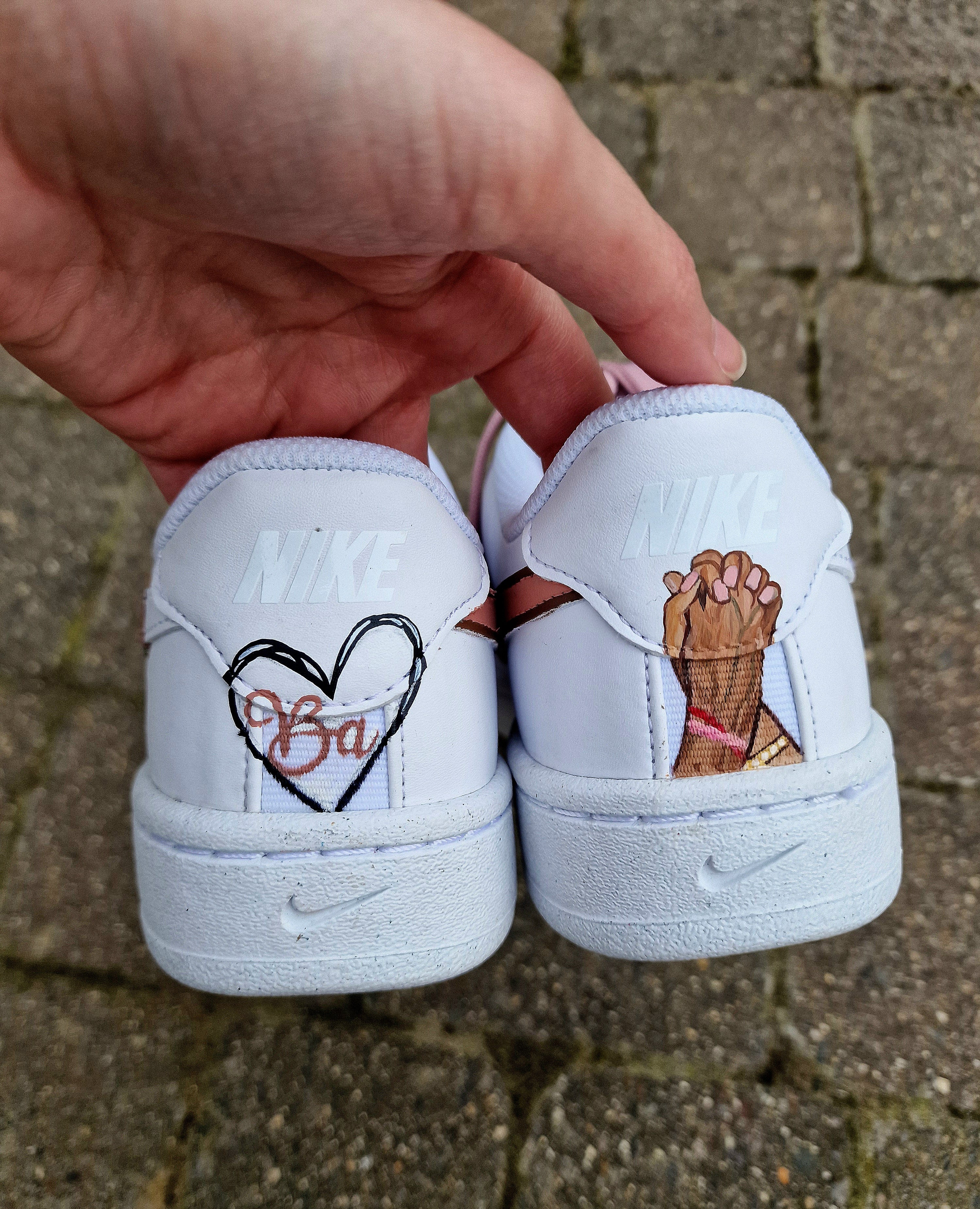 Custom hand-painted Nike sneakers featuring a realistic portrait of a mother and her daughters, personalized names, floral details, and loving elements like “Mama” and “Ba.” Crafted in white leather with pink swooshes and laces, these shoes are a perfect Mother’s Day gift showcasing unique wearable art.

