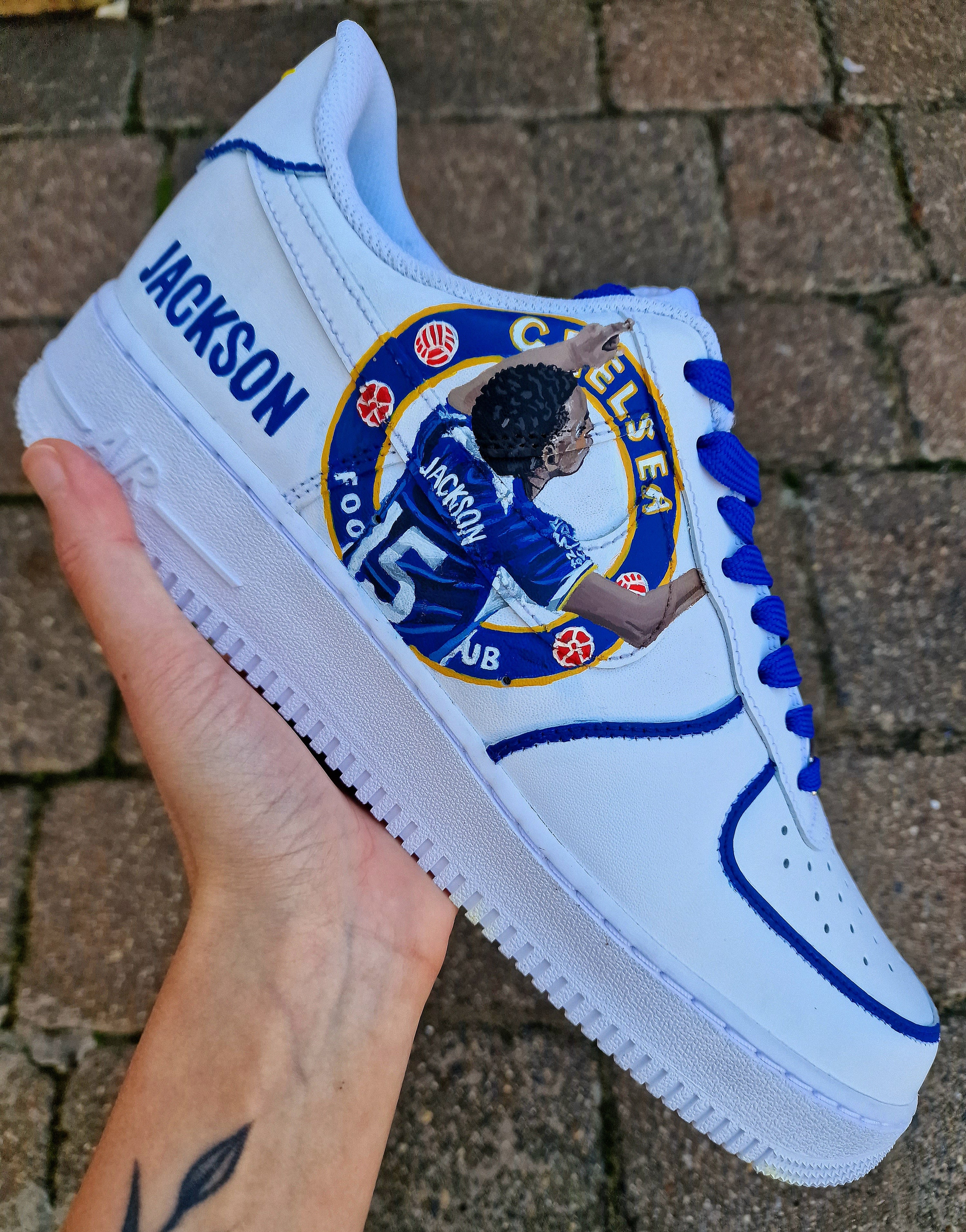 Custom hand-painted white sneakers featuring Chelsea FC’s Nicolas Jackson in vivid blue match kit, complete with the club badge and Senegal flag-inspired Nike swoosh. Designed by Gitartsy, the artwork includes realistic player portraits, fine detailing, and premium waterproof paint, making these shoes a perfect blend of fashion and football art.

