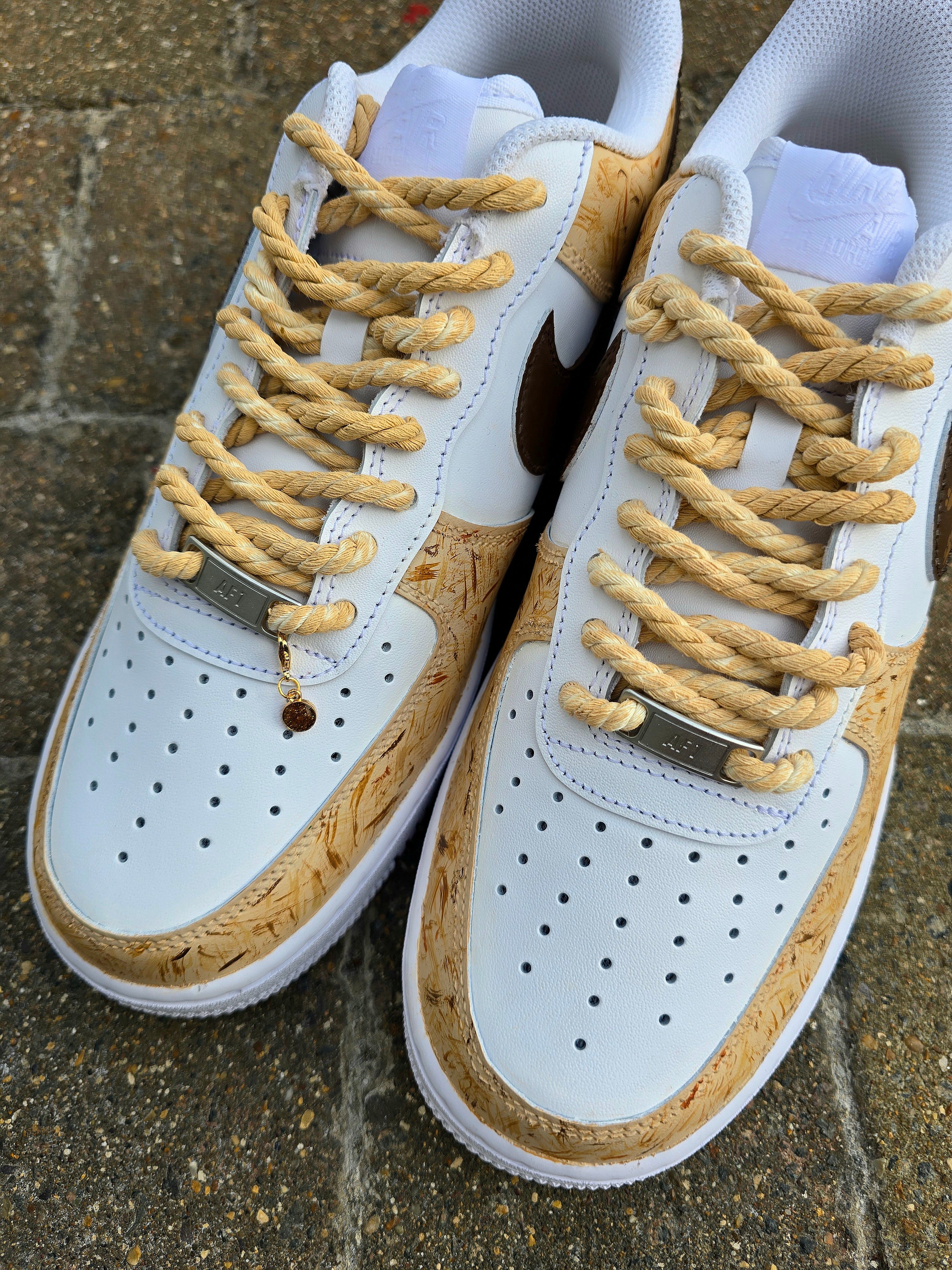 Custom hand-painted Nike Air Force 1 sneakers featuring a detailed portrait of a fluffy brown dog named Poppy 🐶. The shoes have a white leather base with light brown textured paint accents and a rich chocolate-brown swoosh adorned with paw prints 🐾. The pet’s name is written in a playful font, and tan rope laces add a rustic touch. Perfect for showcasing love for your pet in a stylish and artistic way.

