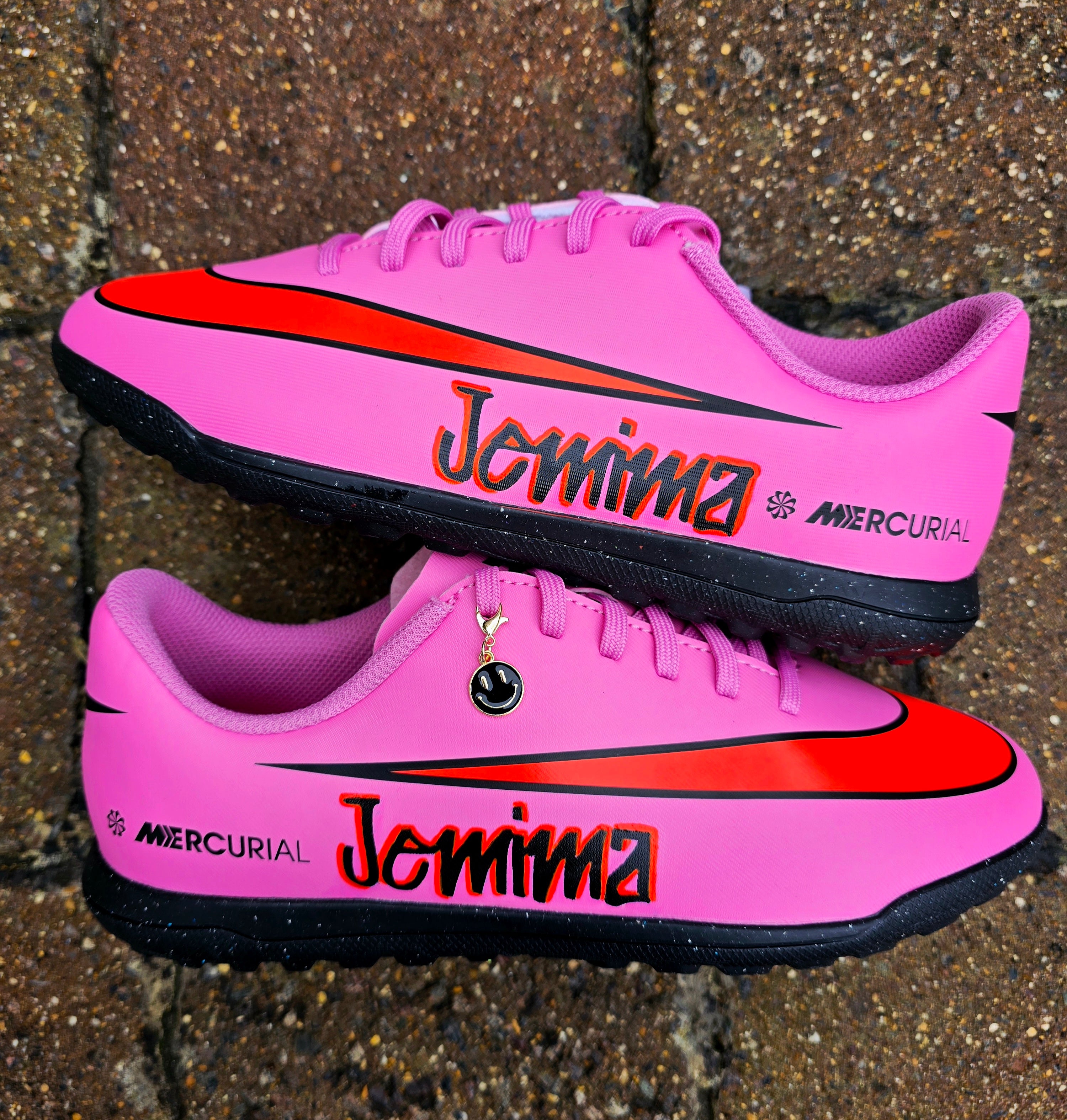 Custom pink Nike Mercurial football sneakers with graffiti-style personalised names in bold red and black lettering. Features include a striking red Nike swoosh, black outsole, and a gold smiley face lace charm. Designed for turf and indoor football, these shoes combine high-performance sportswear with unique, hand-painted street art customisation. Perfect for athletes and collectors seeking one-of-a-kind football boots.

