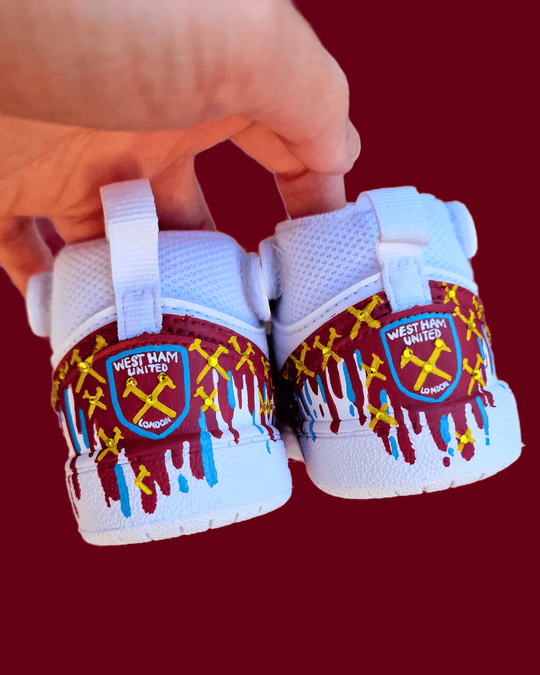 Custom hand-painted baby sneakers inspired by West Ham United, featuring claret and blue color detailing, dripping paint effect, crossed hammers, and personalised names or messages on the straps. Perfect for infant football fans and thoughtful baby gifts.

