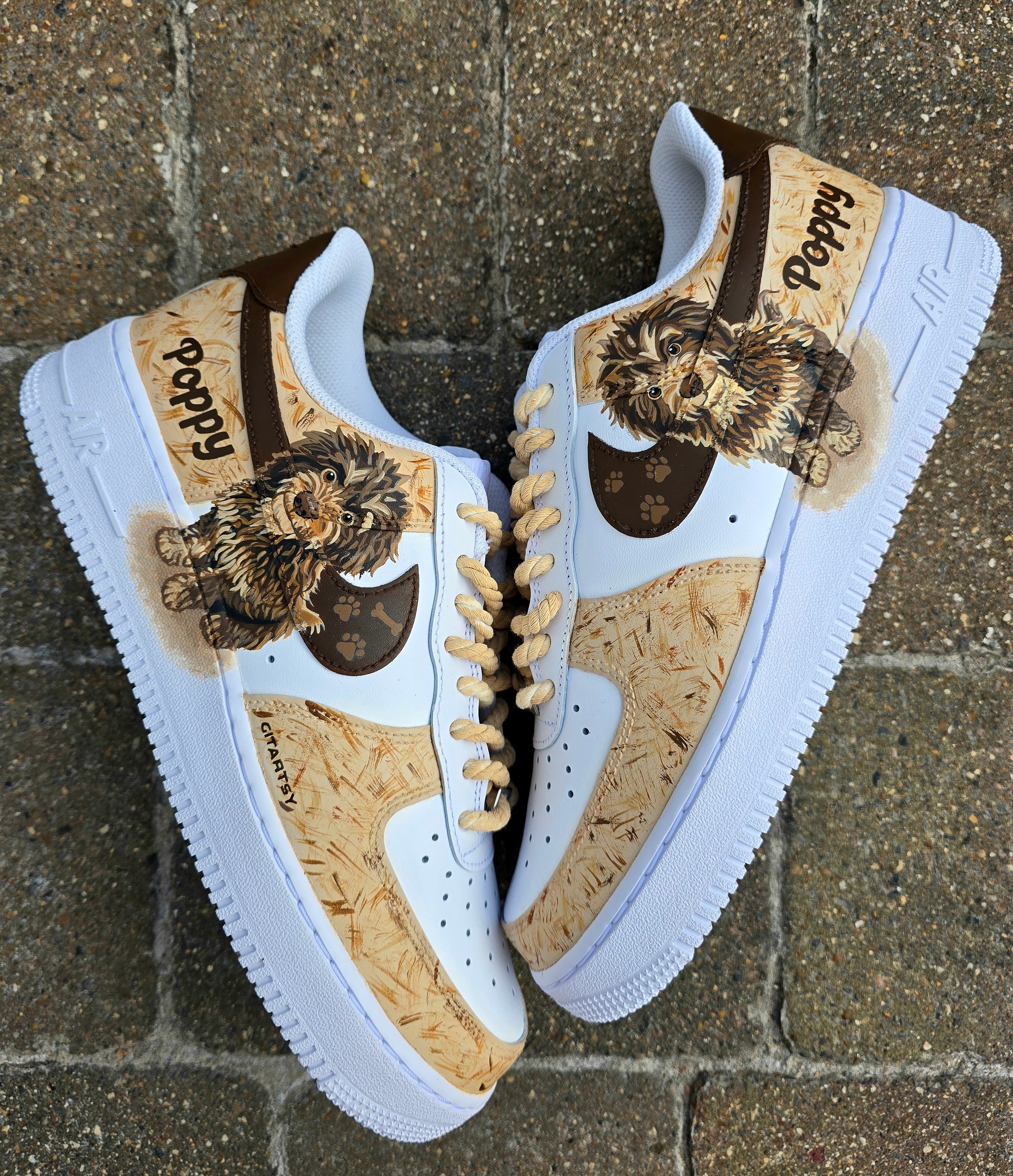 Custom hand-painted Nike Air Force 1 sneakers featuring a detailed portrait of a fluffy brown dog named Poppy 🐶. The shoes have a white leather base with light brown textured paint accents and a rich chocolate-brown swoosh adorned with paw prints 🐾. The pet’s name is written in a playful font, and tan rope laces add a rustic touch. Perfect for showcasing love for your pet in a stylish and artistic way.

