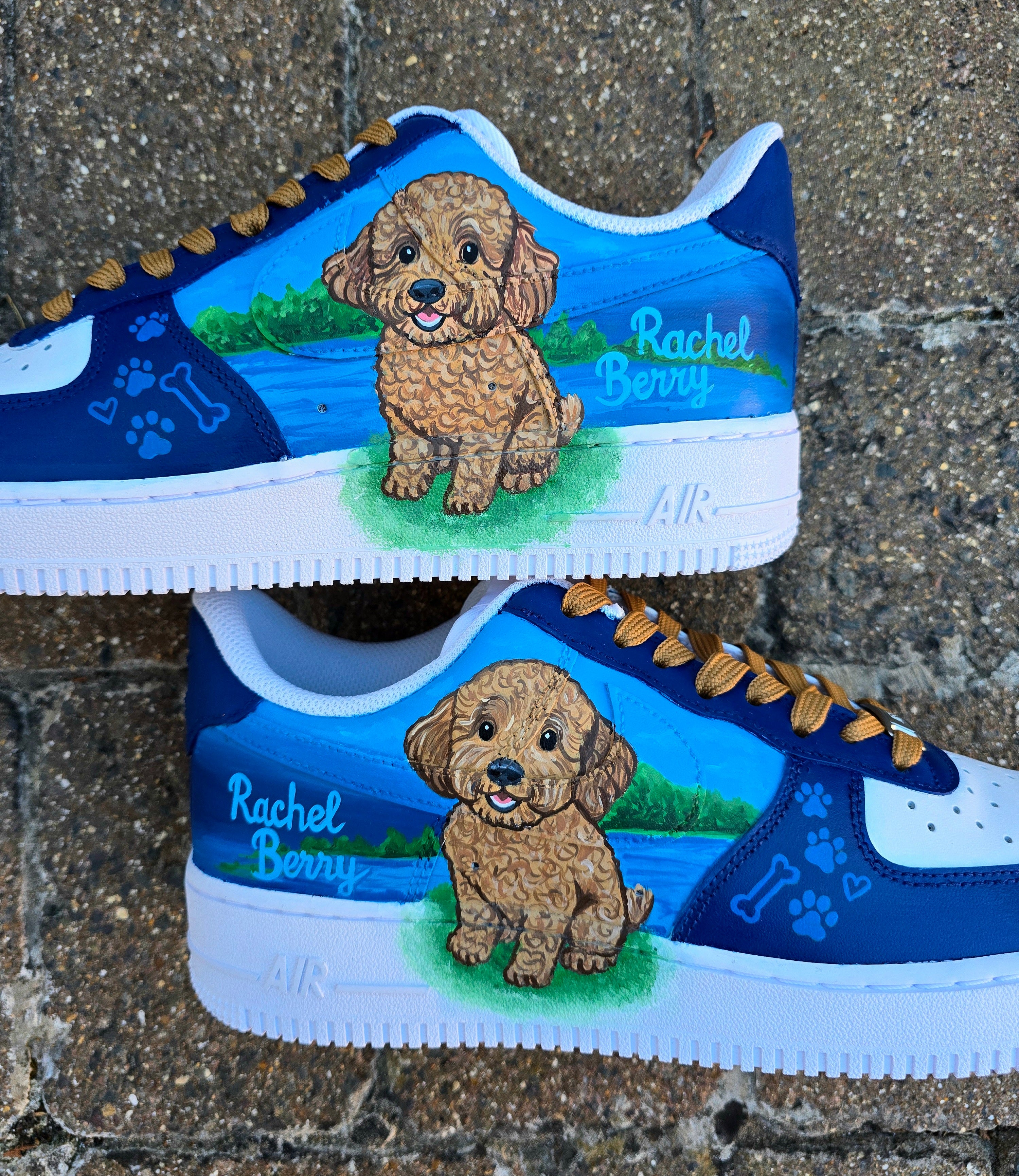 Custom Nike Air Force 1 sneakers 🐾 hand-painted with a detailed toy poodle pet portrait, featuring a caramel-colored curly-haired dog sitting in a grassy field with a blue sky and lake background. Includes the pet’s name in stylish script, paw prints, bones, and heart icons, all painted on a blue and navy color-block base with golden-brown laces. Each pair is personalized to match your pet’s likeness and crafted using durable, waterproof leather paints, making them wearable art for dog lovers.

