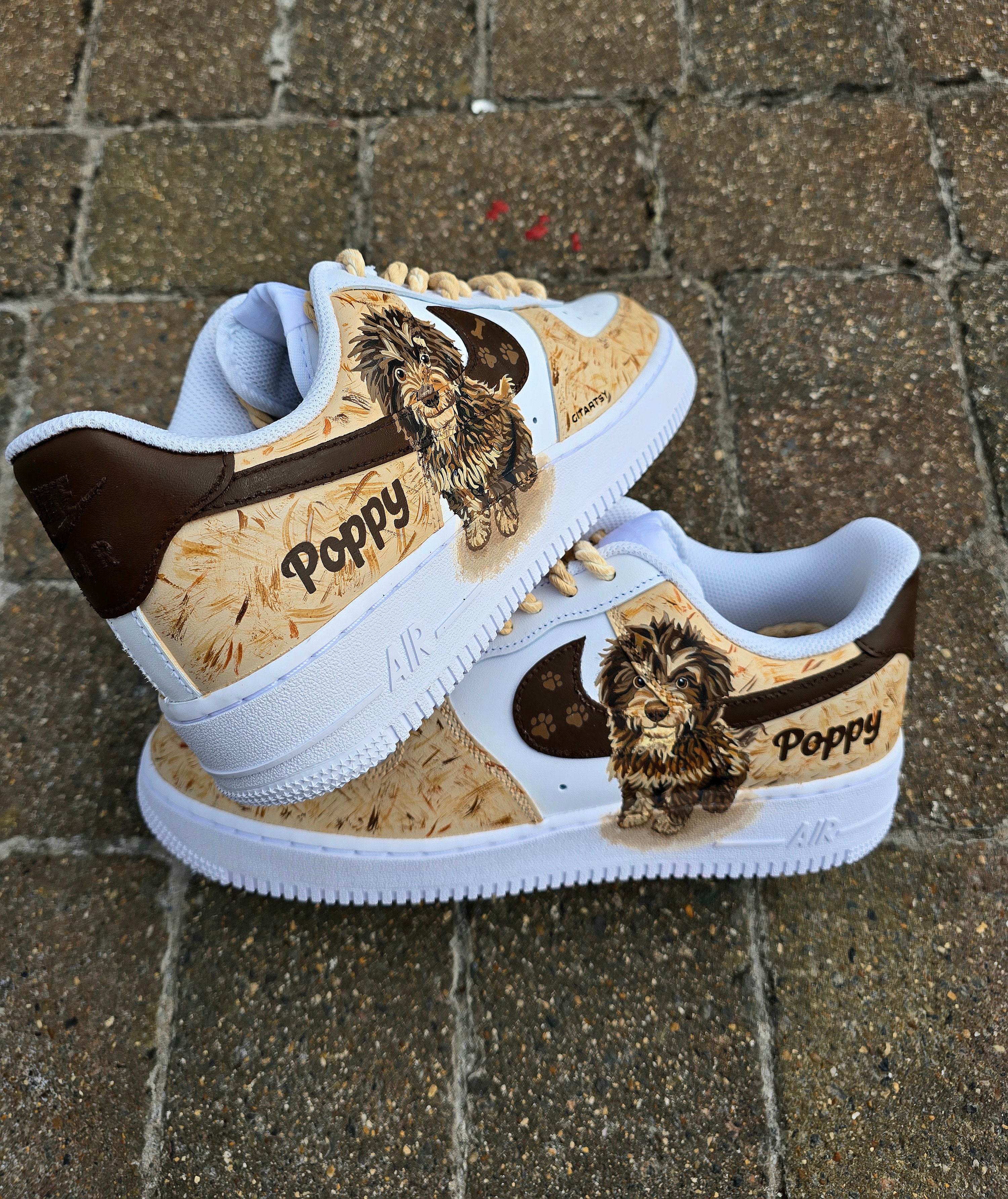 Custom hand-painted Nike Air Force 1 sneakers featuring a detailed portrait of a fluffy brown dog named Poppy 🐶. The shoes have a white leather base with light brown textured paint accents and a rich chocolate-brown swoosh adorned with paw prints 🐾. The pet’s name is written in a playful font, and tan rope laces add a rustic touch. Perfect for showcasing love for your pet in a stylish and artistic way.

