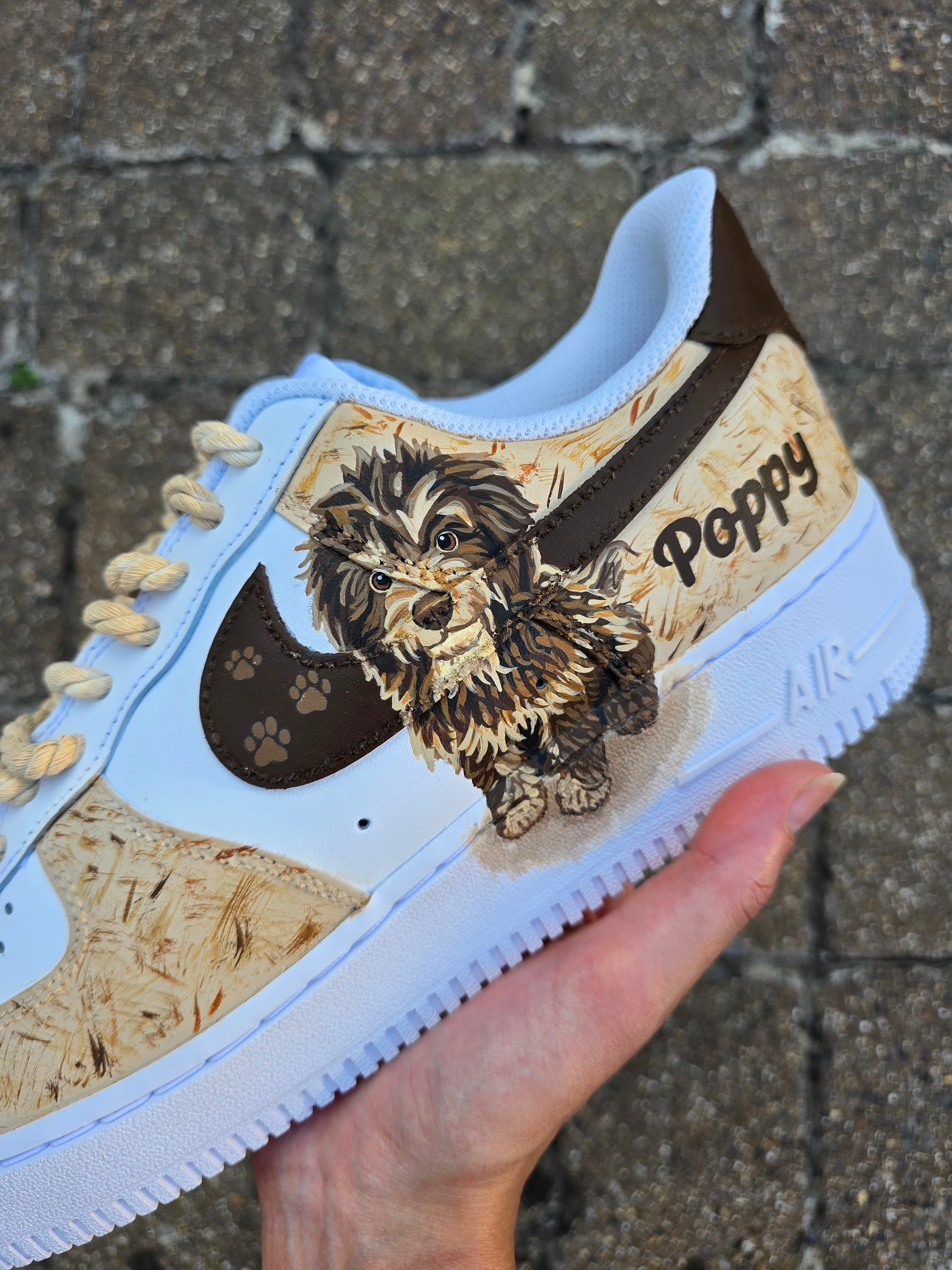 Custom hand-painted Nike Air Force 1 sneakers featuring a detailed portrait of a fluffy brown dog named Poppy 🐶. The shoes have a white leather base with light brown textured paint accents and a rich chocolate-brown swoosh adorned with paw prints 🐾. The pet’s name is written in a playful font, and tan rope laces add a rustic touch. Perfect for showcasing love for your pet in a stylish and artistic way.

