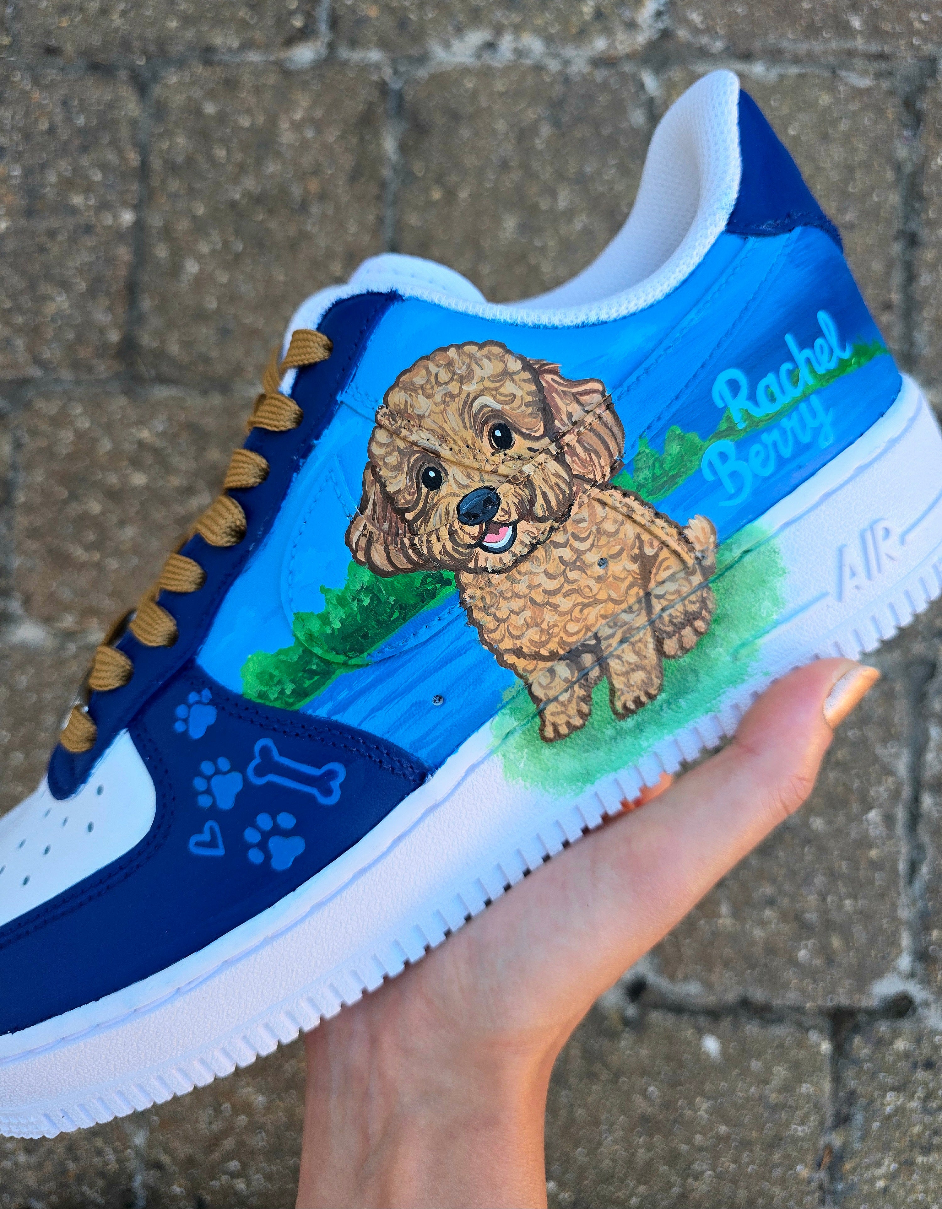 Custom Nike Air Force 1 sneakers 🐾 hand-painted with a detailed toy poodle pet portrait, featuring a caramel-colored curly-haired dog sitting in a grassy field with a blue sky and lake background. Includes the pet’s name in stylish script, paw prints, bones, and heart icons, all painted on a blue and navy color-block base with golden-brown laces. Each pair is personalized to match your pet’s likeness and crafted using durable, waterproof leather paints, making them wearable art for dog lovers.

