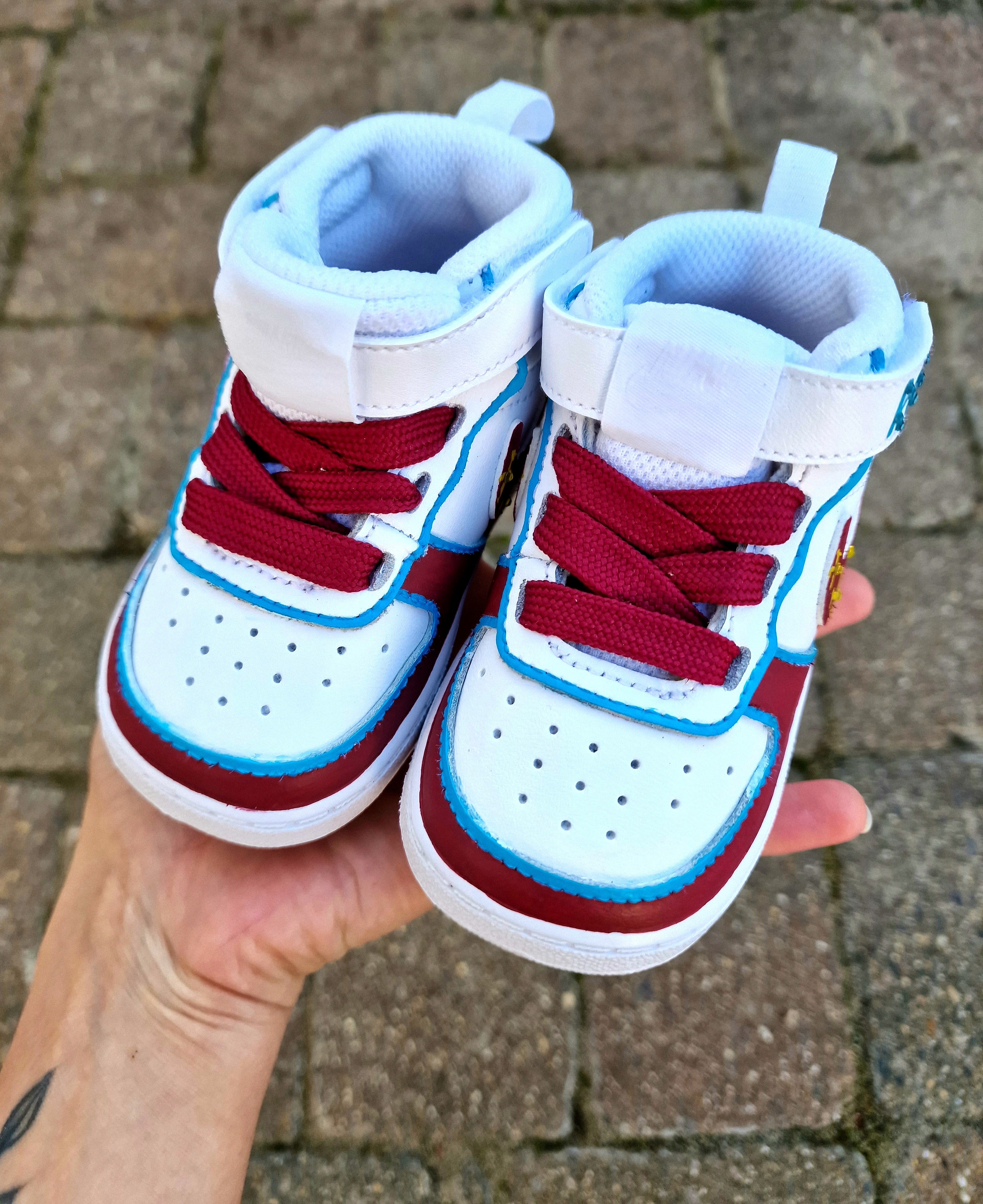 Custom hand-painted baby sneakers inspired by West Ham United, featuring claret and blue color detailing, dripping paint effect, crossed hammers, and personalised names or messages on the straps. Perfect for infant football fans and thoughtful baby gifts.

